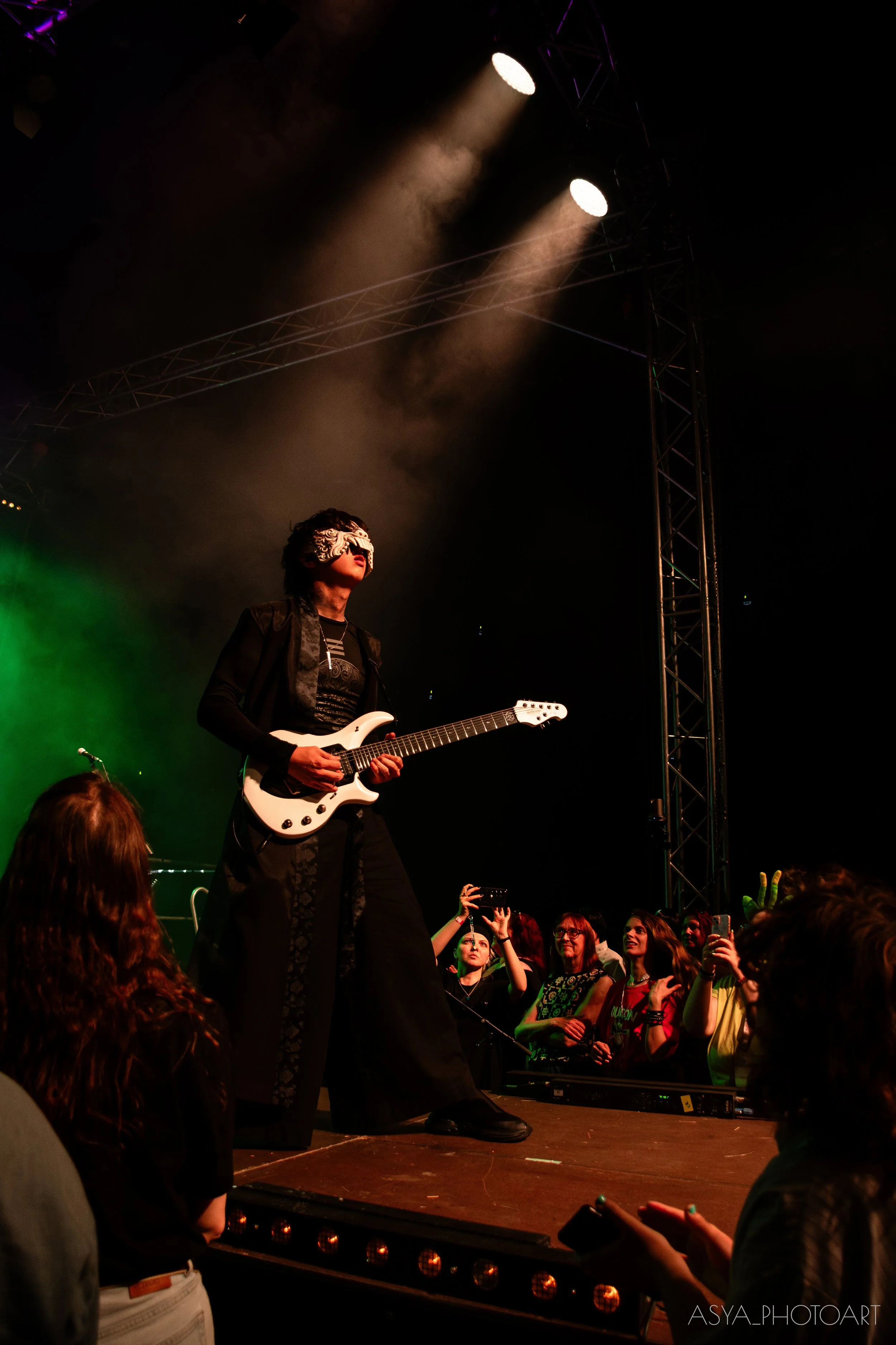 A musician playing an electric guitar on stage during a concert, with a crowd watching in the foreground. The stage is lit with bright spotlights and colored lights, creating a vibrant atmosphere.