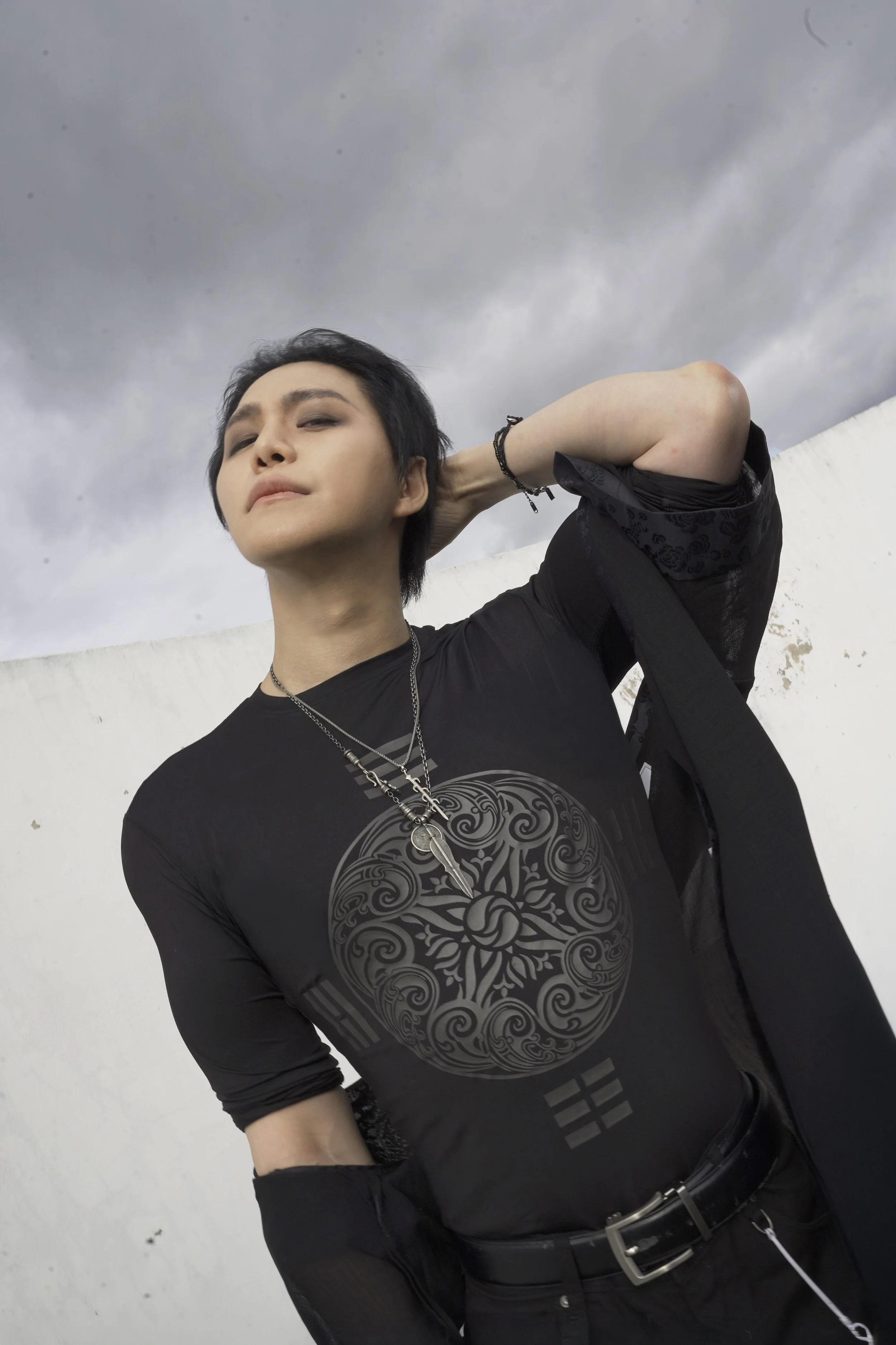 A person with short black hair, wearing a black shirt with a detailed circular design on the front, standing outdoors against a cloudy sky, looking into the distance with a thoughtful expression, with one hand behind their head and the other hand in 