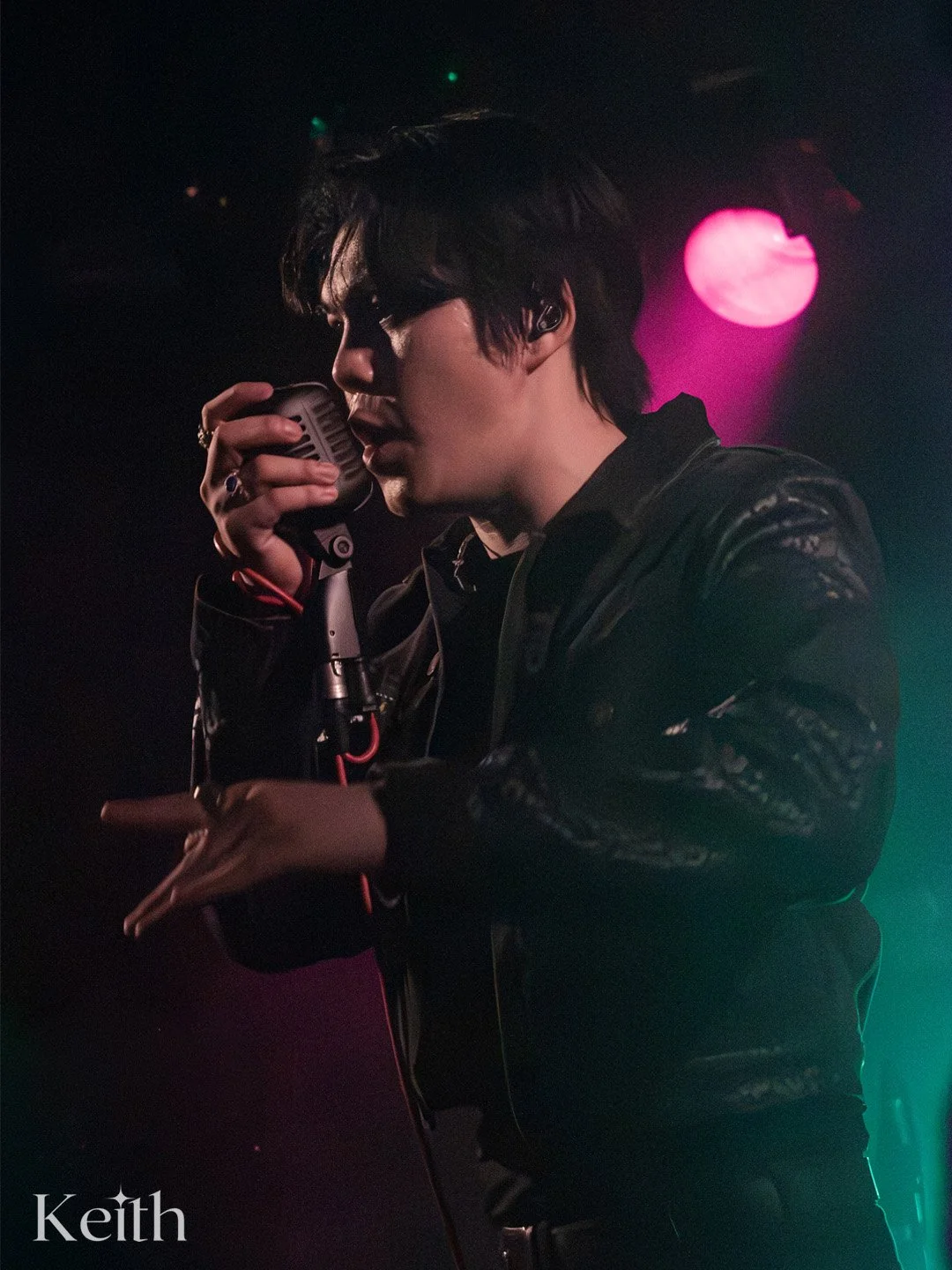 A male musician singing into a microphone on stage, illuminated by pink and green stage lights, wearing a black jacket.