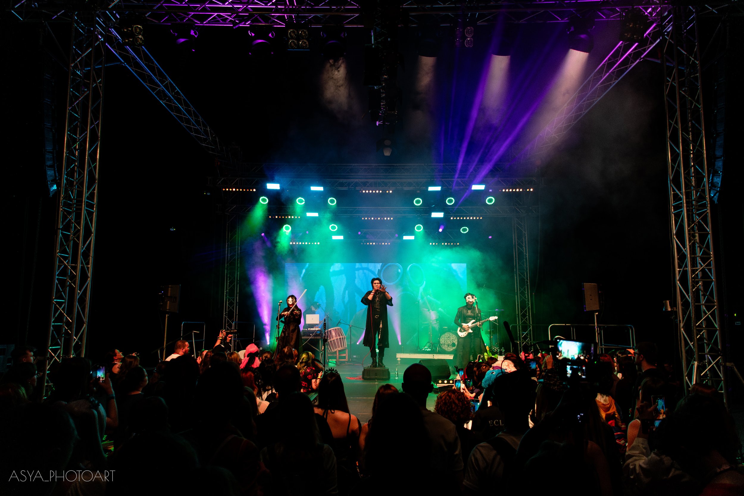 A live music concert on a stage with colorful lighting, featuring a singer, a violinist, and a guitarist, with an audience in front of the stage.