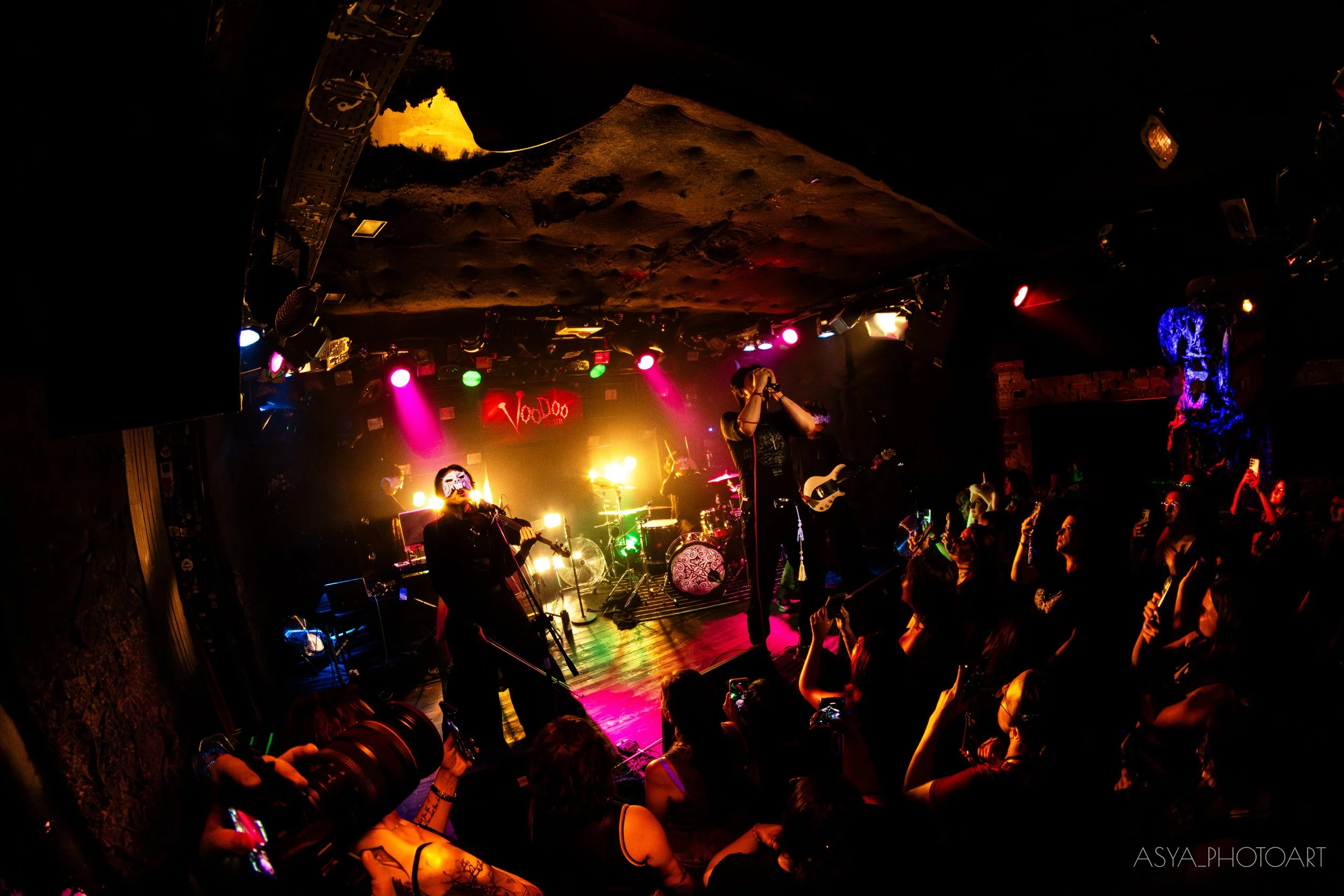 A live music performance on a stage with colorful lighting, a singer with a microphone, musicians playing instruments, and an audience watching, in a dark venue.