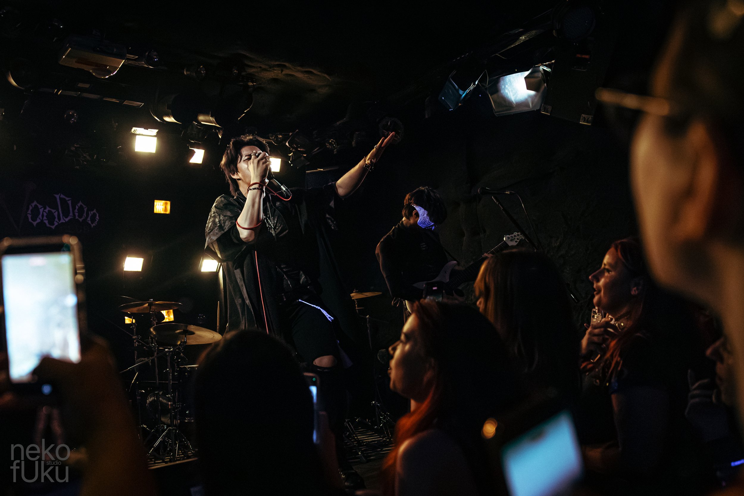 A live music performance in a dark venue with a singer holding a microphone and reaching out to the audience, surrounded by band members and fans taking photos.