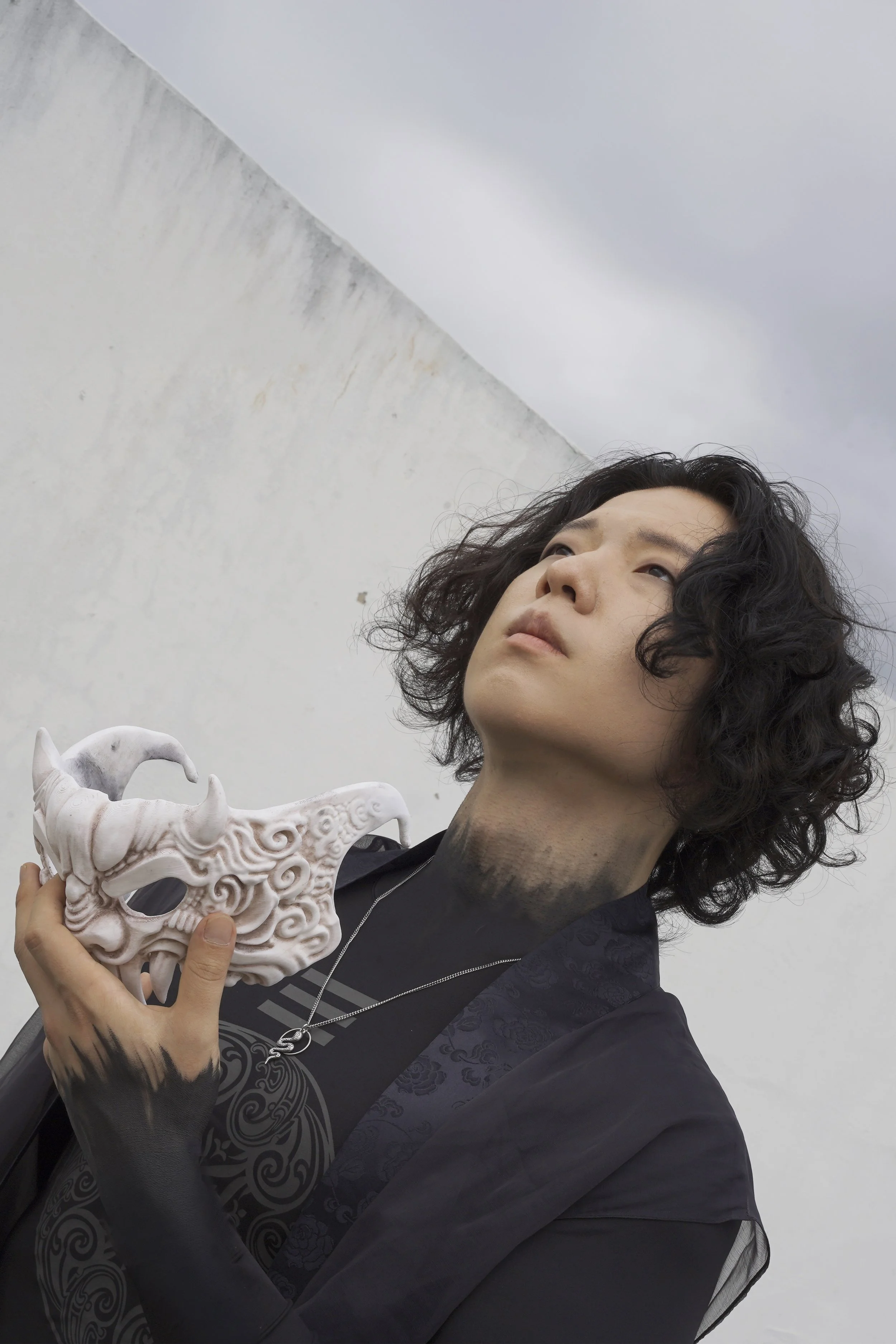 A person with curly dark hair wearing a black shirt with a swirling design, holding a white ornate mask resembling a dragon or mythical creature, standing against a plain gray wall.
