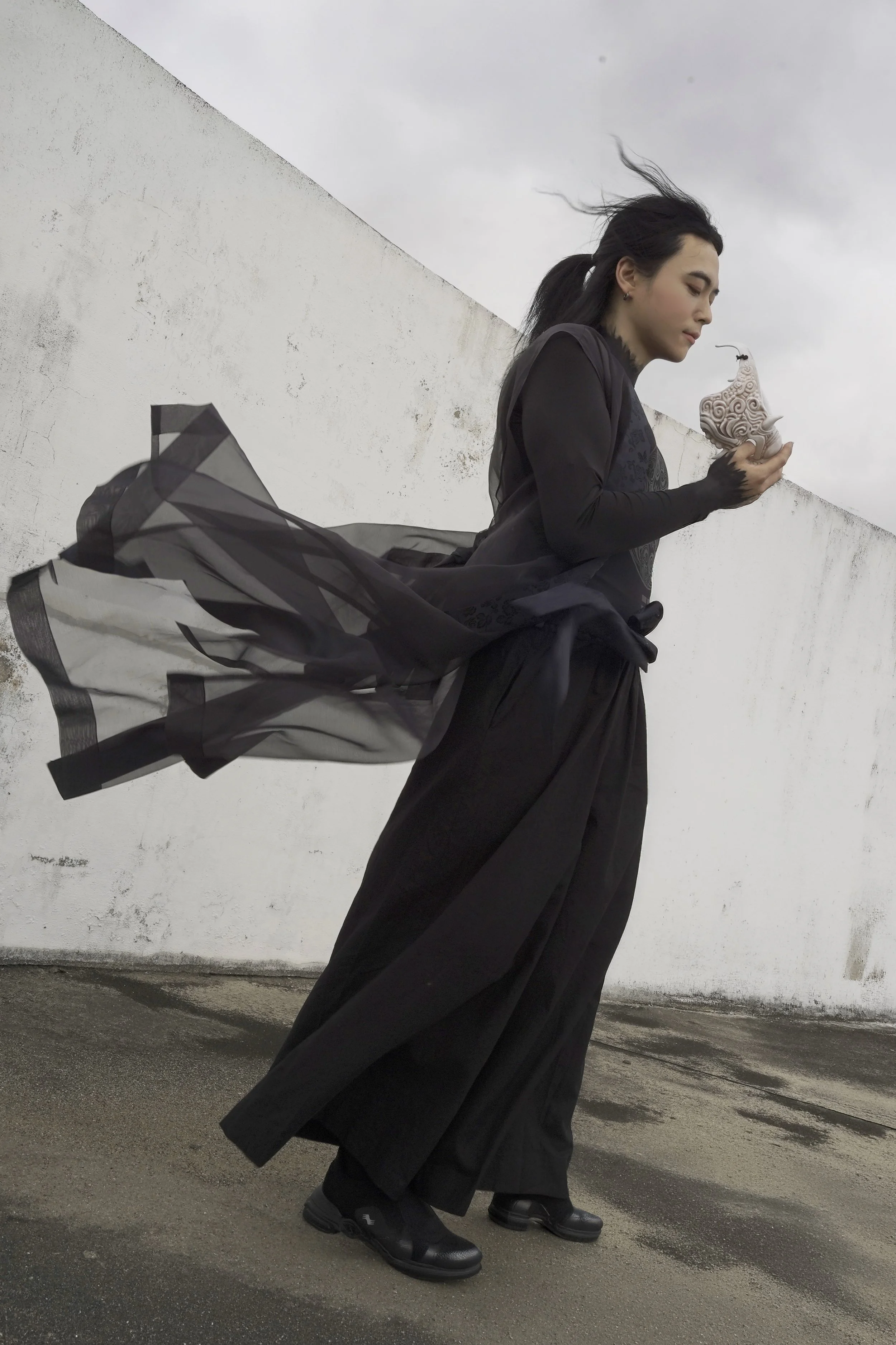 A woman dressed in black standing against a white wall, holding a decorative candle holder with a candle, and wearing black boots and a flowing black skirt.