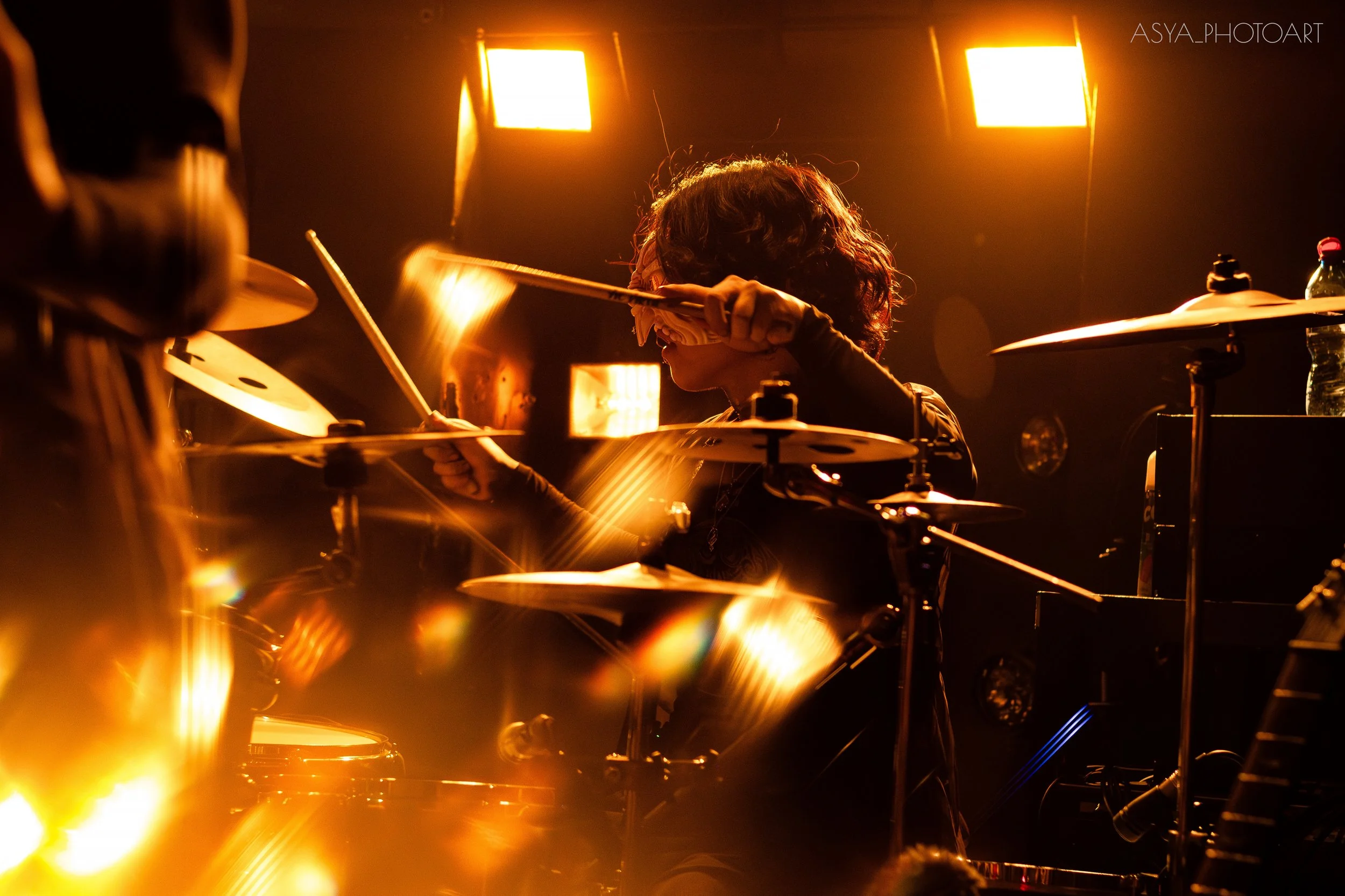 Person with curly hair playing drums in a dark setting with orange stage lights.