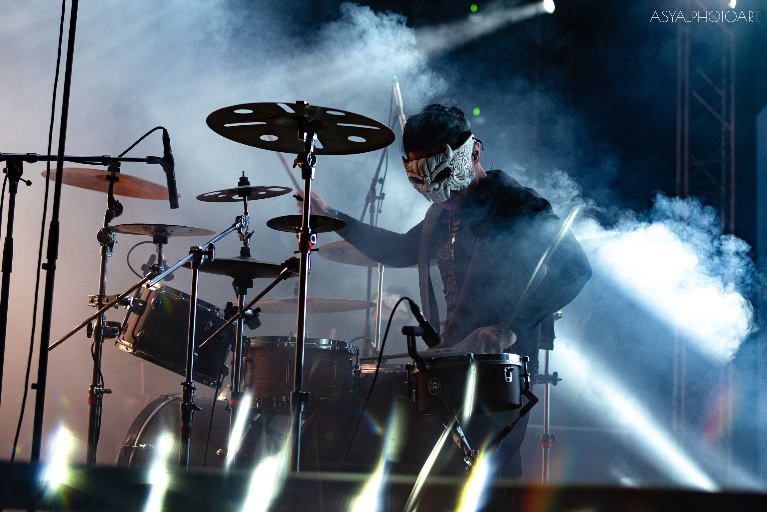 A musician with a demon mask playing drums on stage surrounded by stage smoke and intense lighting.