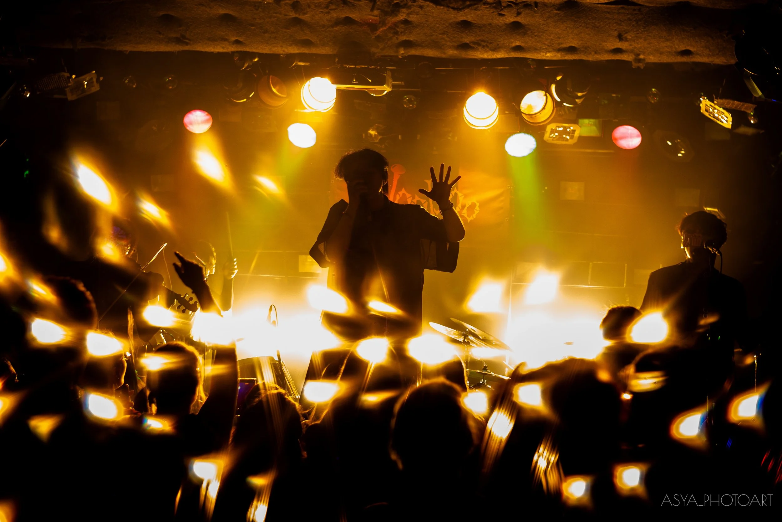 A person on stage with colorful stage lights shining behind them, surrounded by an audience and visible instruments.