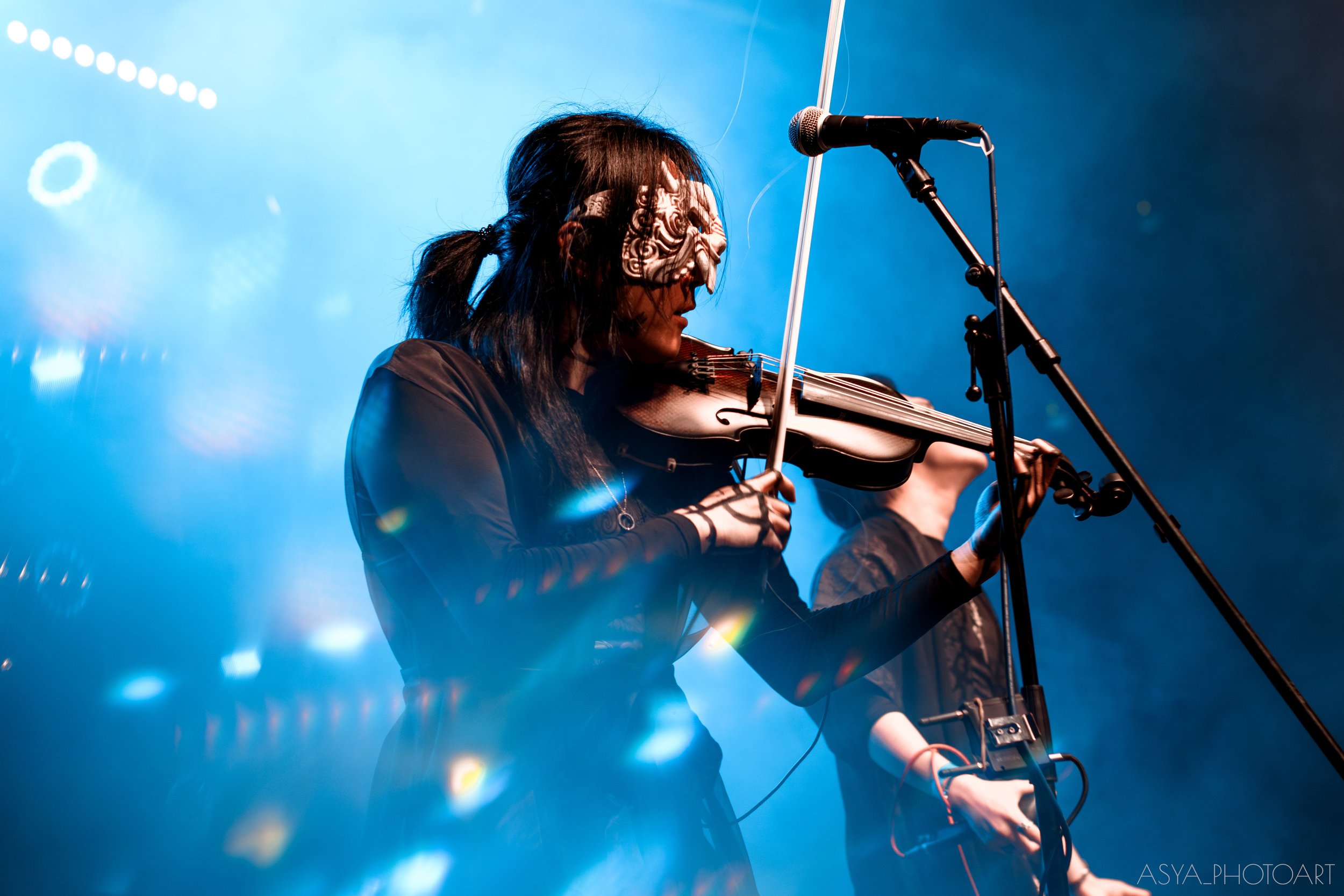 A woman with long dark hair playing a violin on stage, wearing a decorative mask over her eyes, with blue stage lighting and a microphone in front of her.