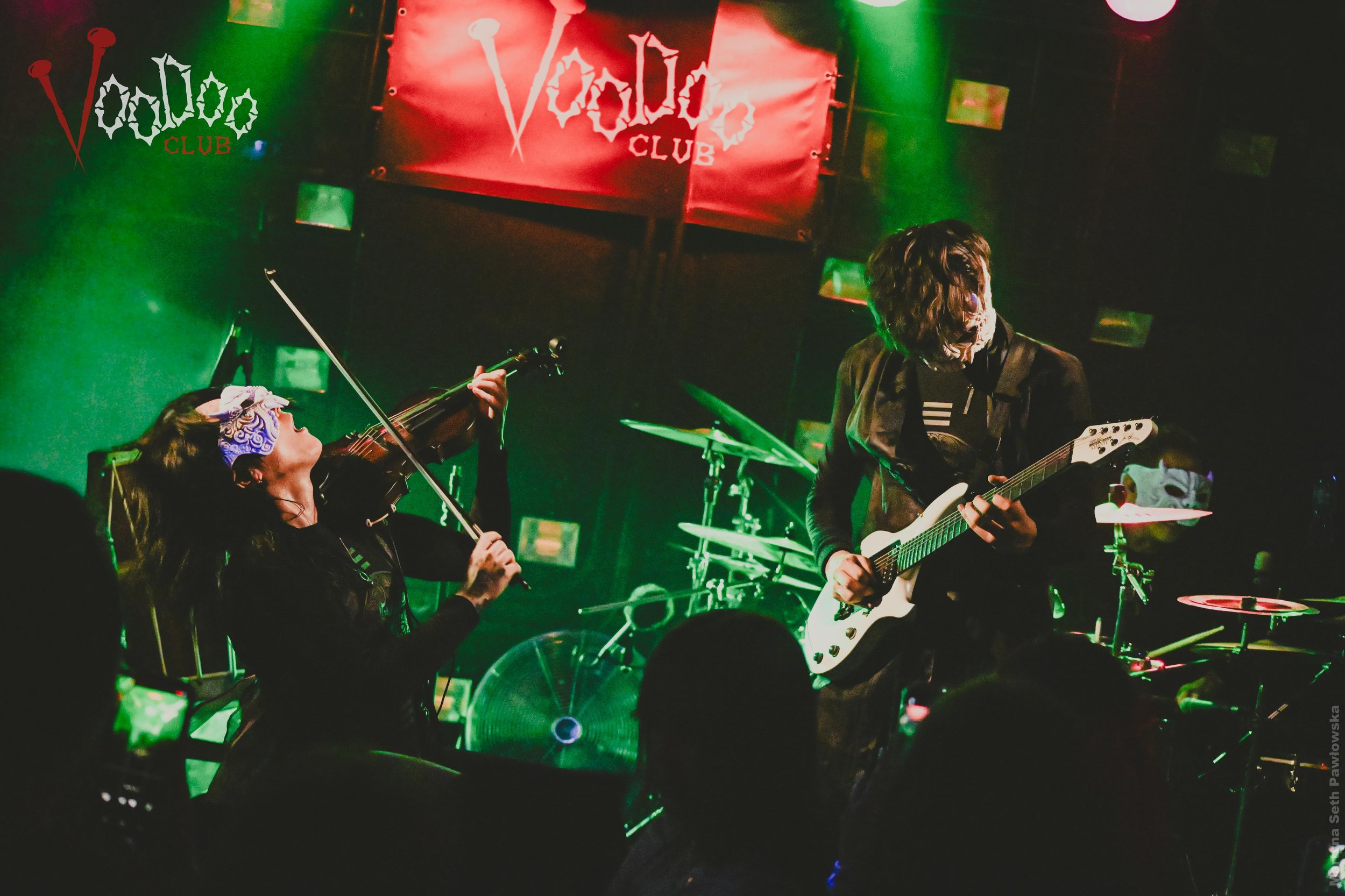 A live music performance at VooDoo Club with a female violinist and a male guitarist on stage, with a red and green lighting setup and a red club sign in the background.
