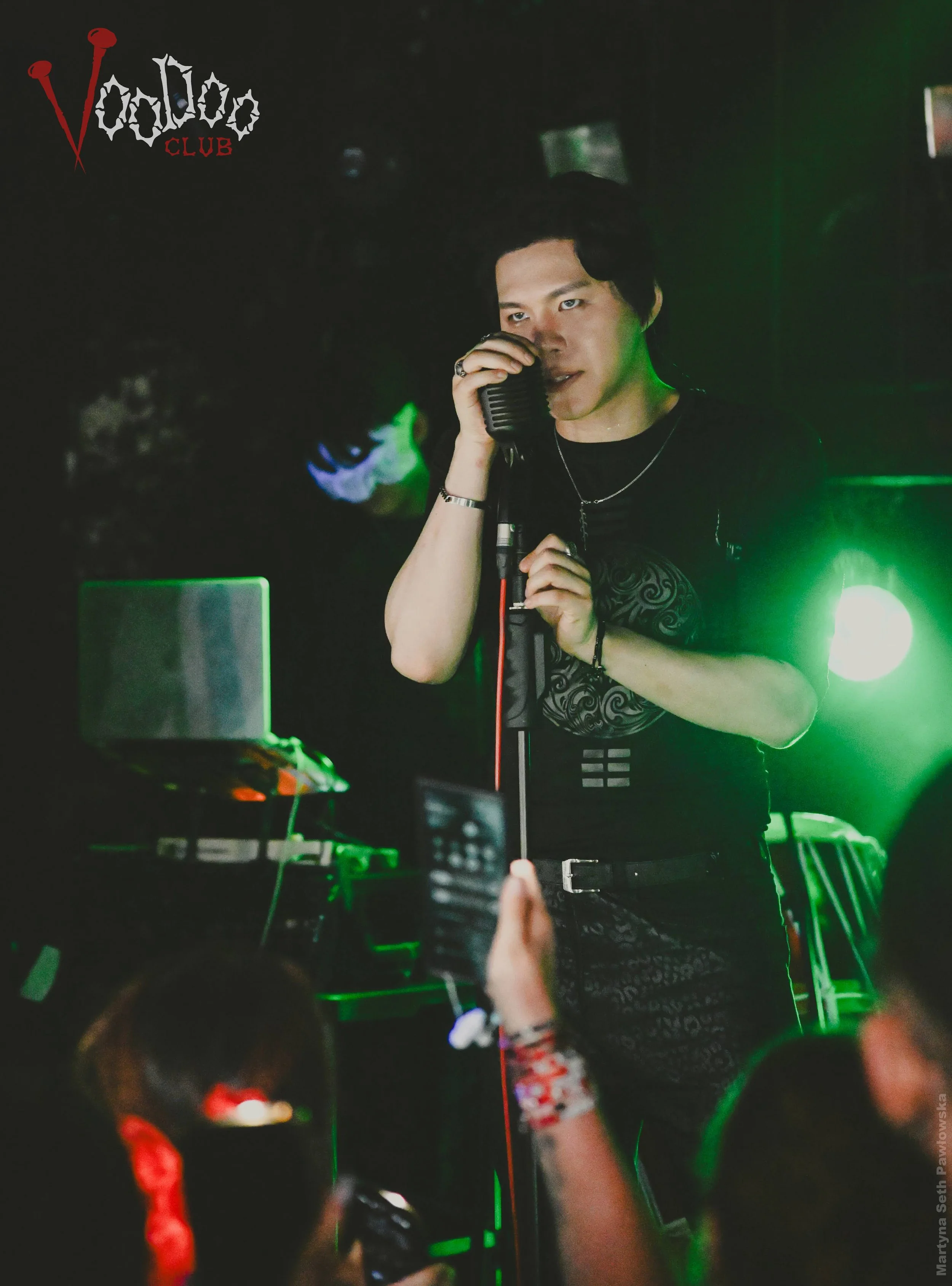 A person with dark hair singing into a vintage microphone in a dark club with green lighting, with a logo reading 'Voodoo CLUB' in the top left corner.