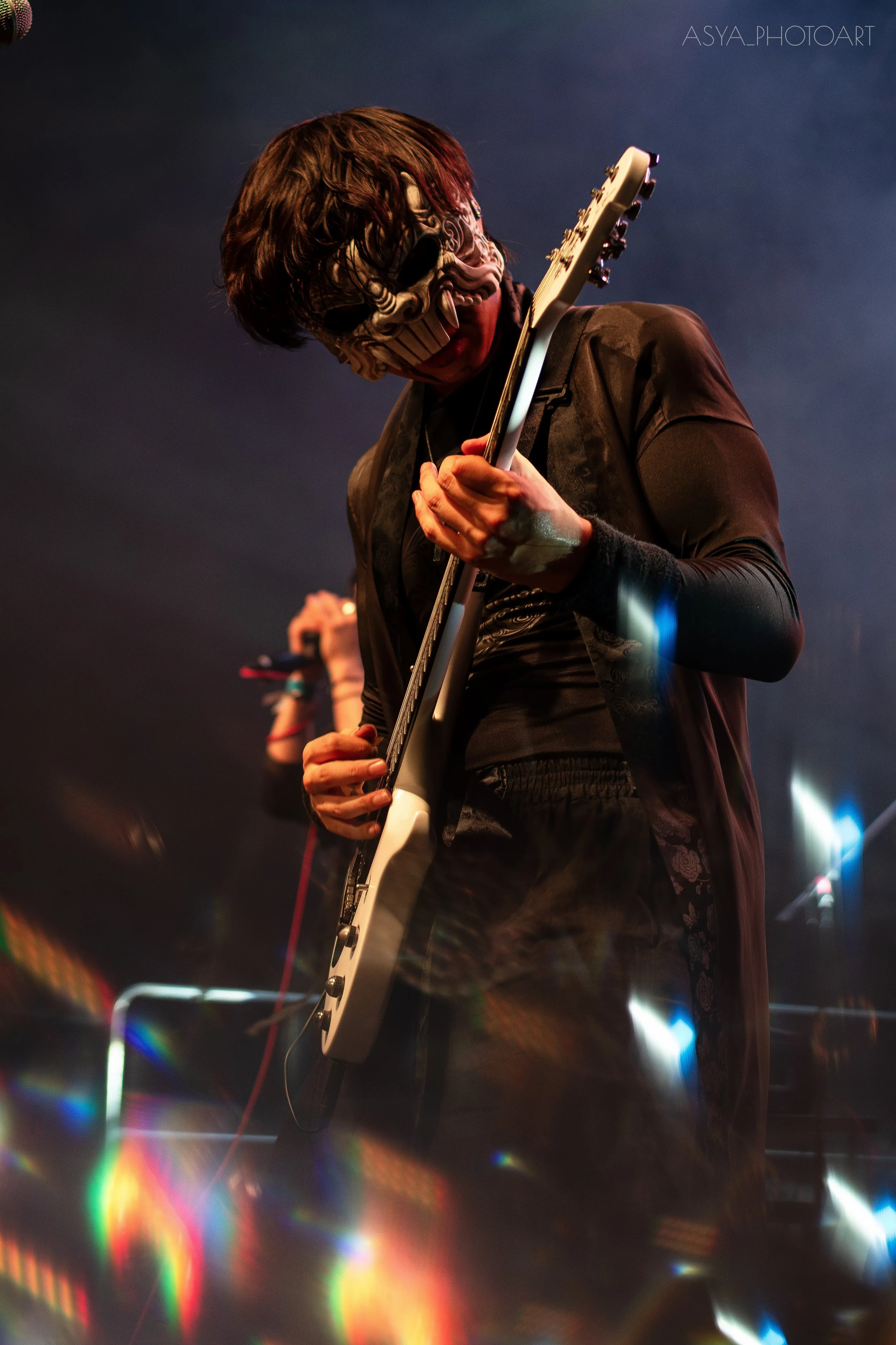 Person playing an electric guitar at a concert, wearing a skeleton mask and dark clothing, with spotlights and colorful light effects in the background.