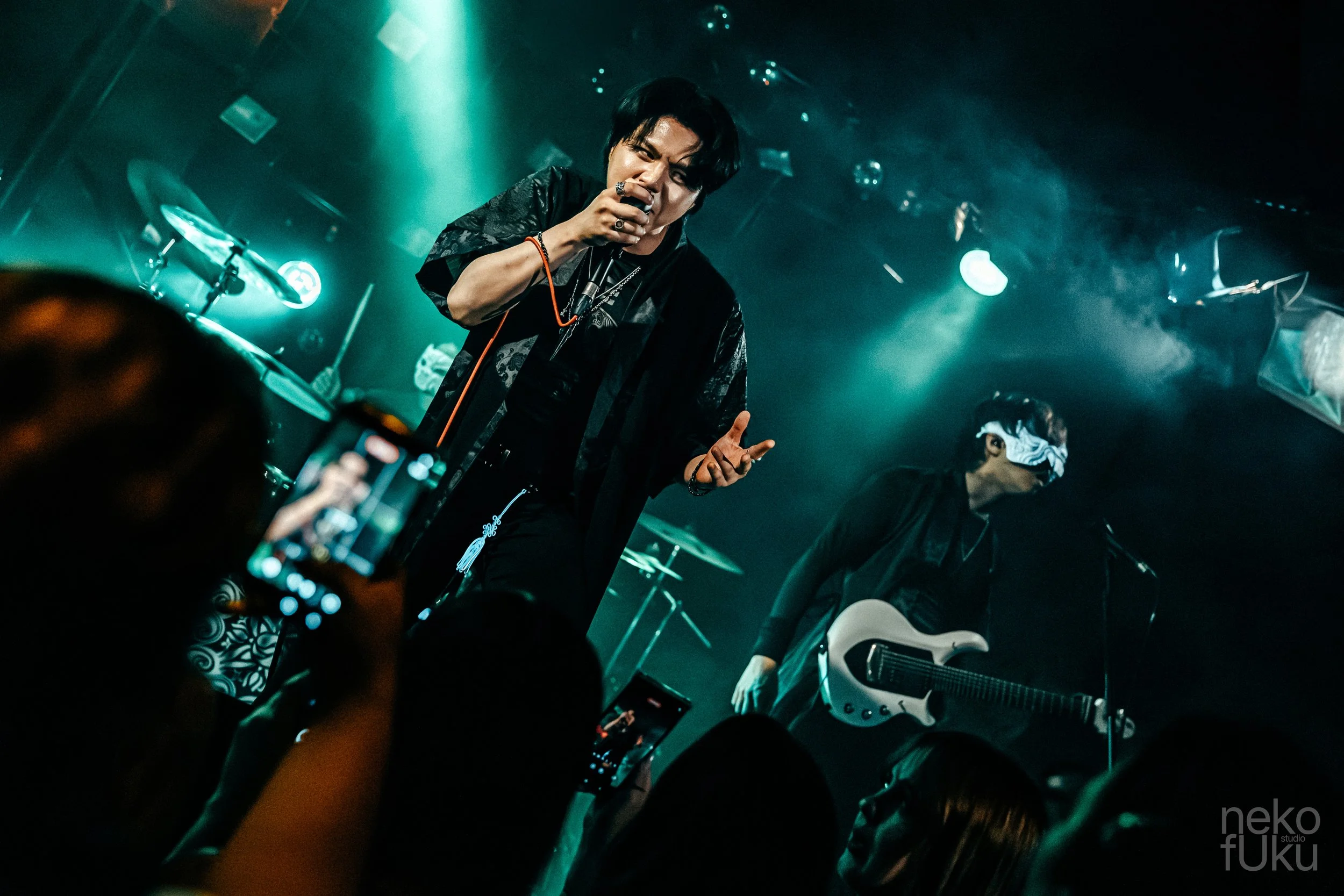 A male singer with black hair performing on stage, holding a microphone, with other band members and musical instruments visible in a dimly lit setting with blue and green stage lights.