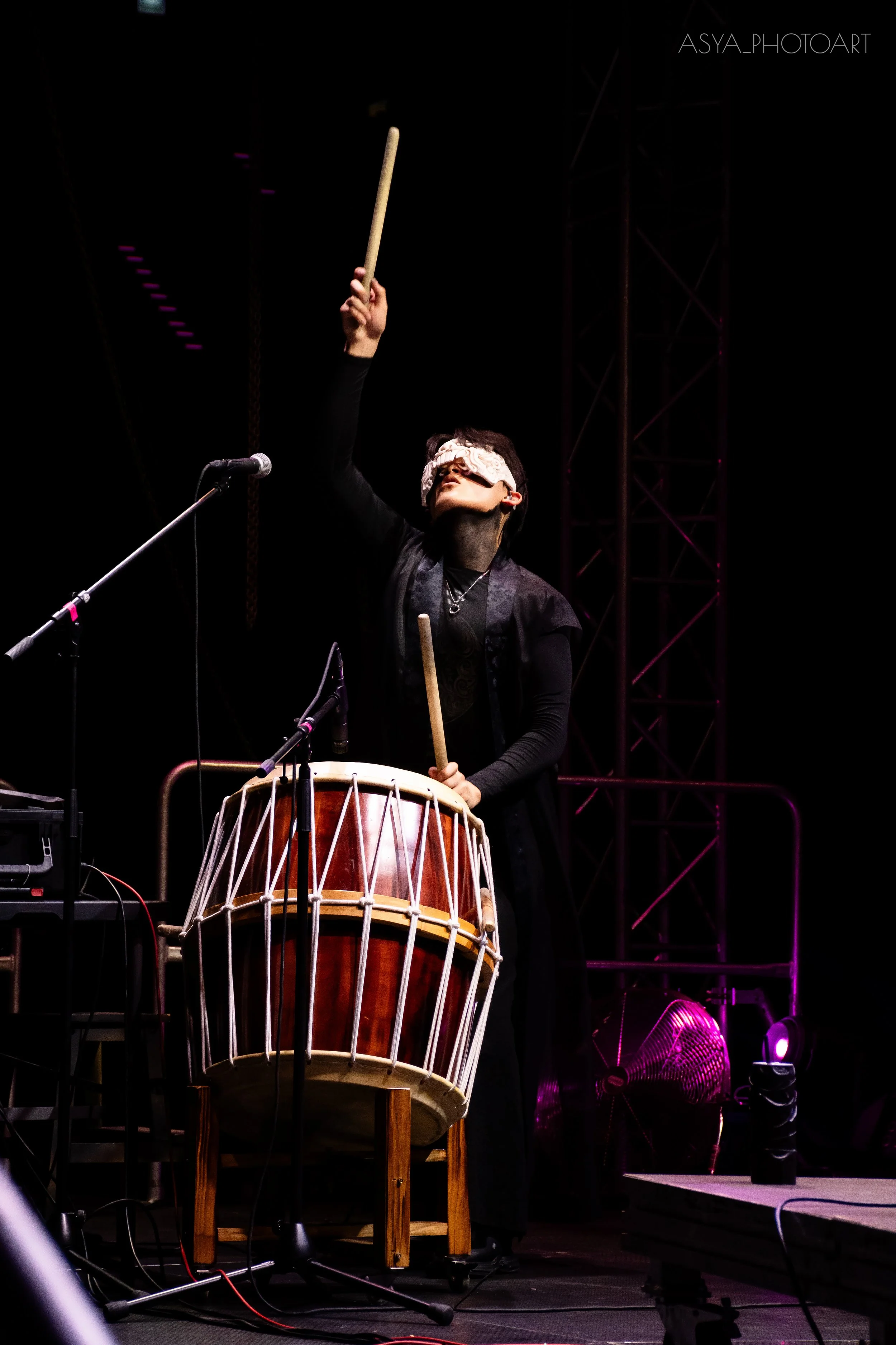 A performer is playing a traditional drum on stage, wearing an eye mask and holding a drumstick raised in the air. Bright stage lighting highlights the performer and drum, with a microphone set up nearby.