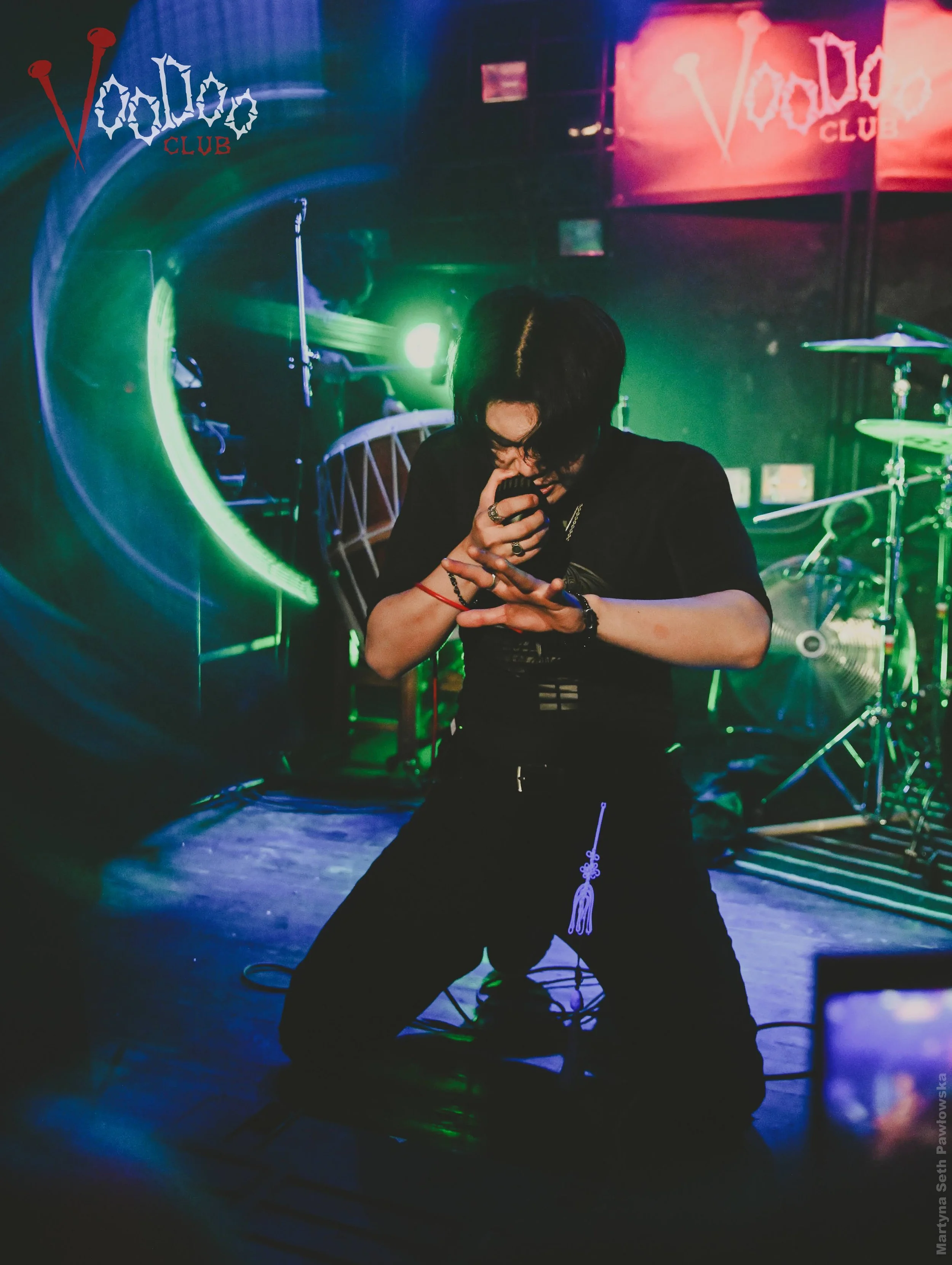 Person with black hair and rings singing into a microphone on stage in a rock music venue called Voodoo Club, with drums and neon lights visible in the background.