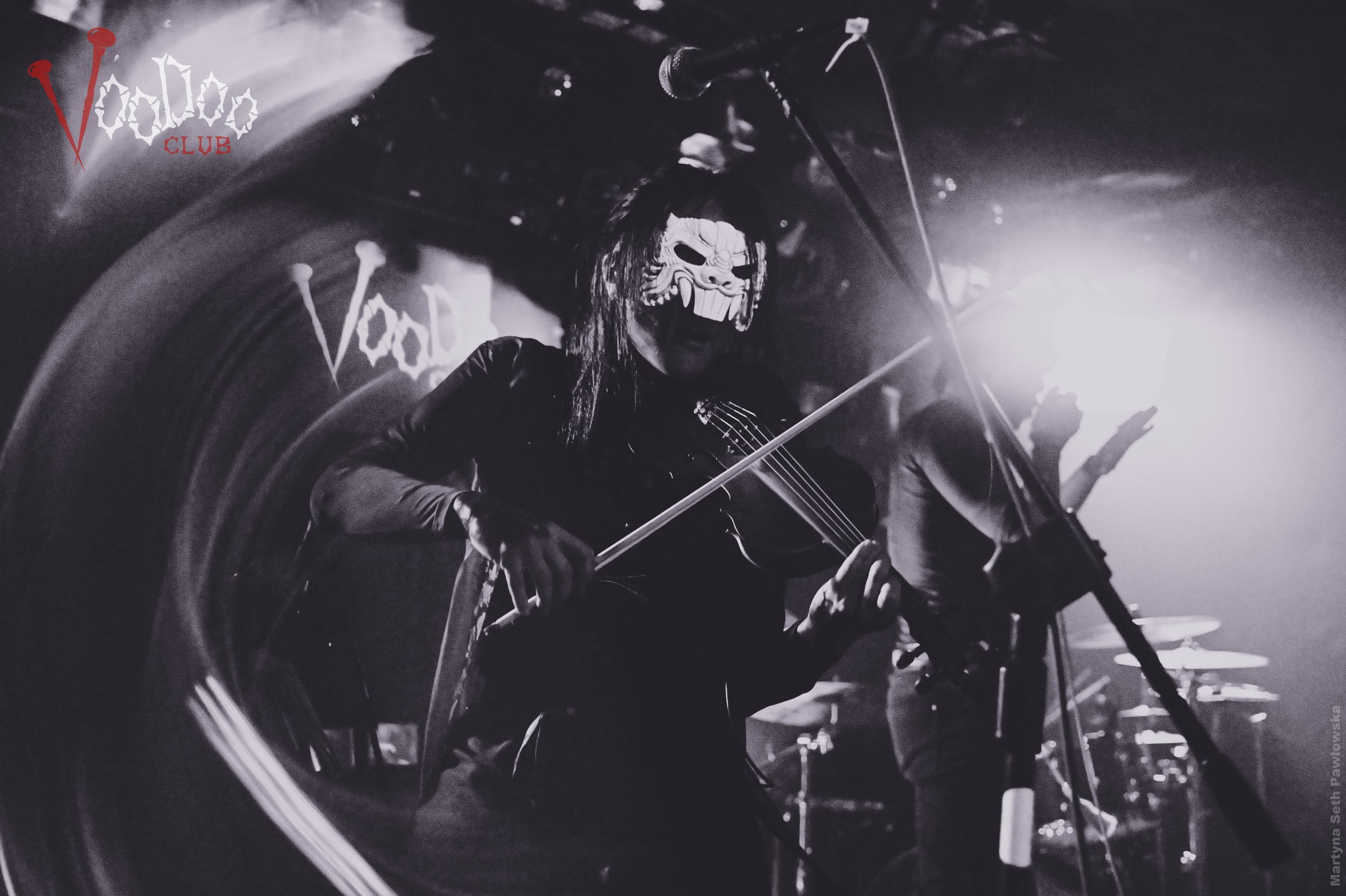 A musician wearing a skeleton mask is playing a keyboard on stage, with other band members in the background. The scene is in black and white, with a illuminated logo that reads 'Voodoo Club' in the top left corner.