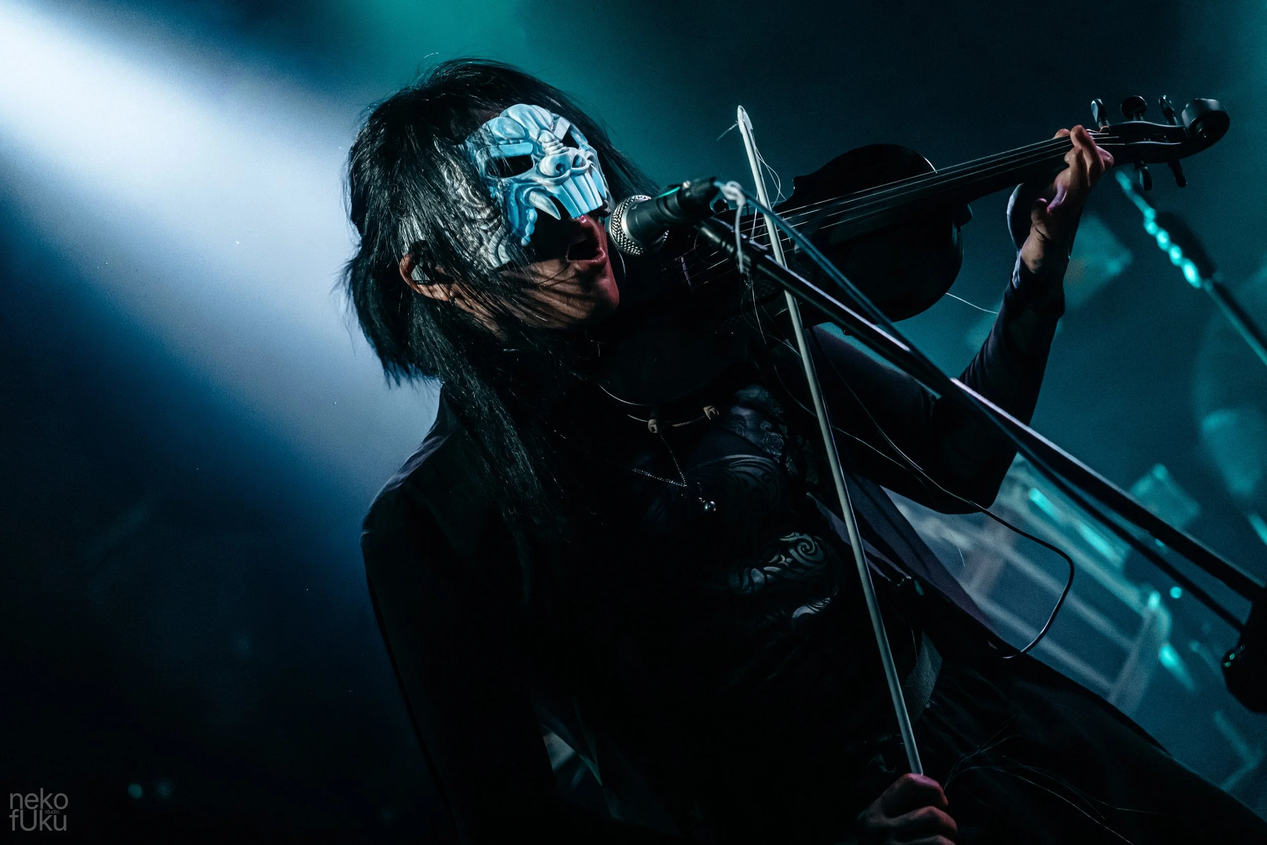 A performer wearing a blue mask with a fierce expression, black clothing, long black hair, playing a guitar on stage with bright stage lights and a foggy atmosphere.