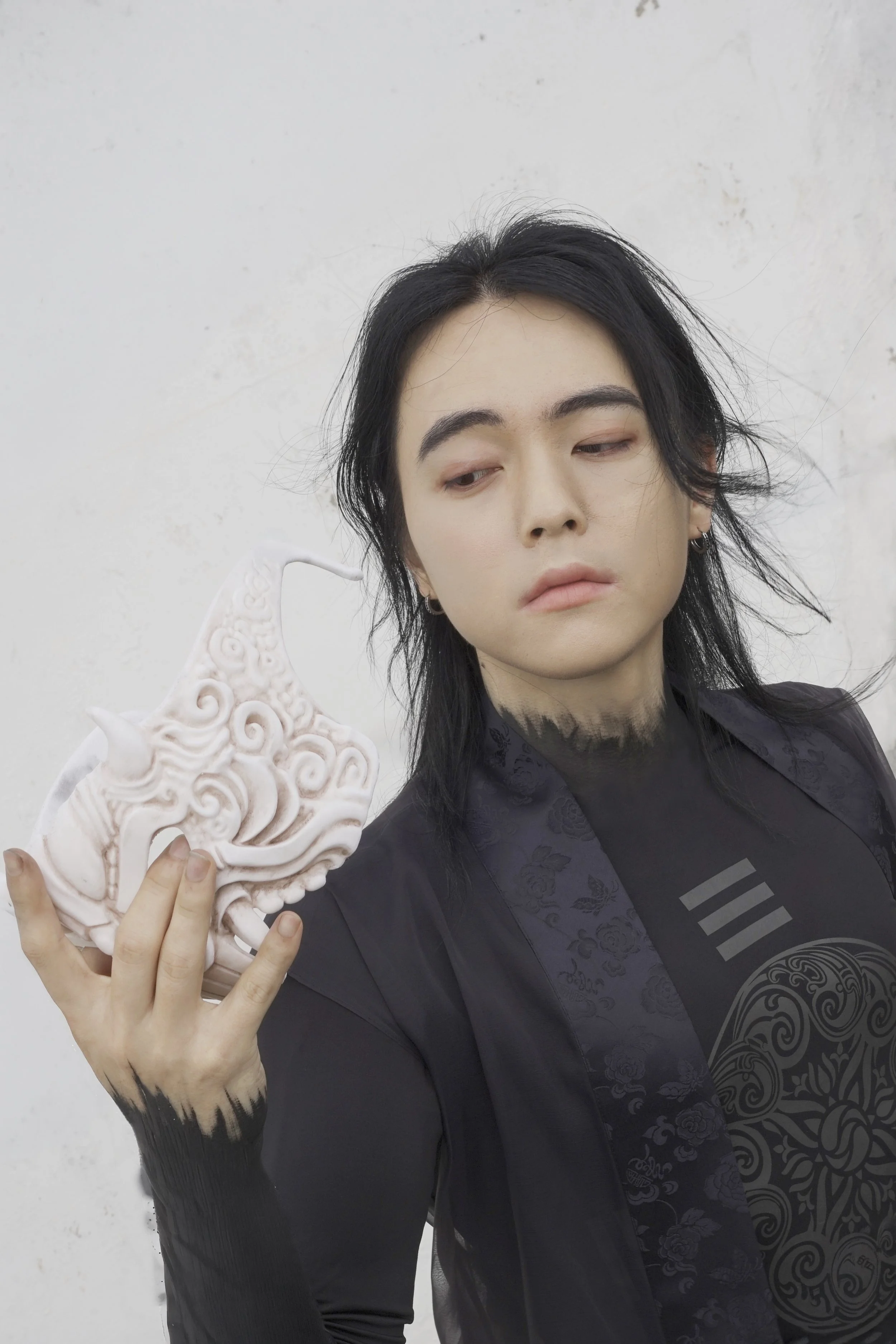 A person with black hair, wearing a dark jacket with Asian-inspired patterns, holding a decorative white ceramic sculpture of a Chinese dragon with swirling clouds, against a plain white wall.