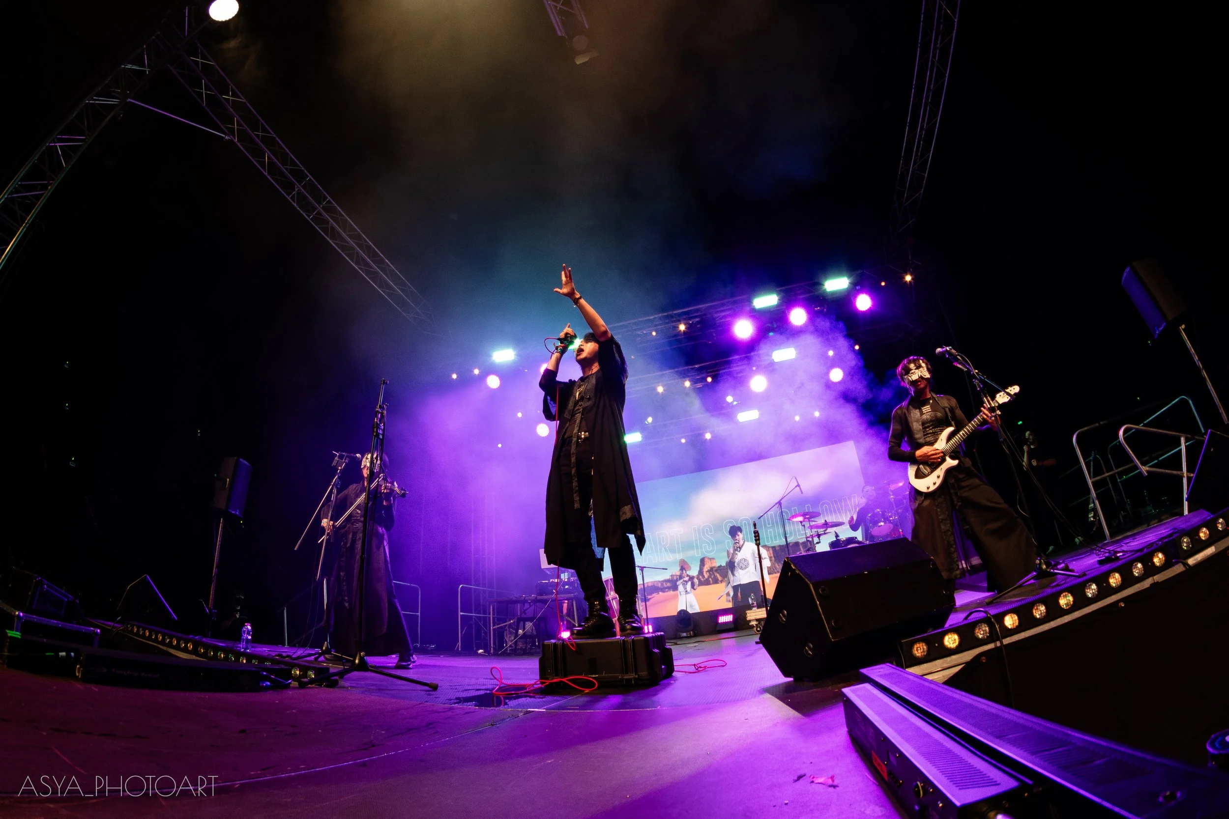 A band performs on stage at night with colorful purple, white, and blue lighting. The lead singer is in the center holding a microphone, and another musician is playing a guitar on the right. The stage has various equipment and large screens in the b