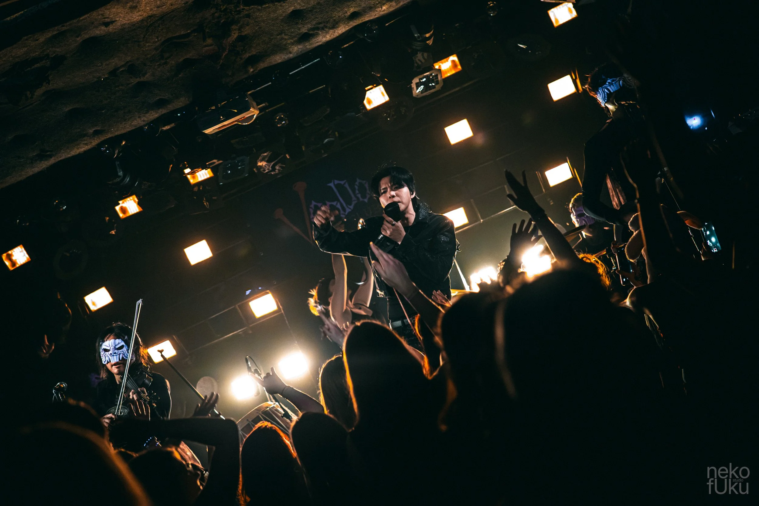 A live concert with a singer holding a microphone on stage, surrounded by audience members with raised hands. The stage is dimly lit with bright orange and white lights and features rock or metal band members, some wearing masks.