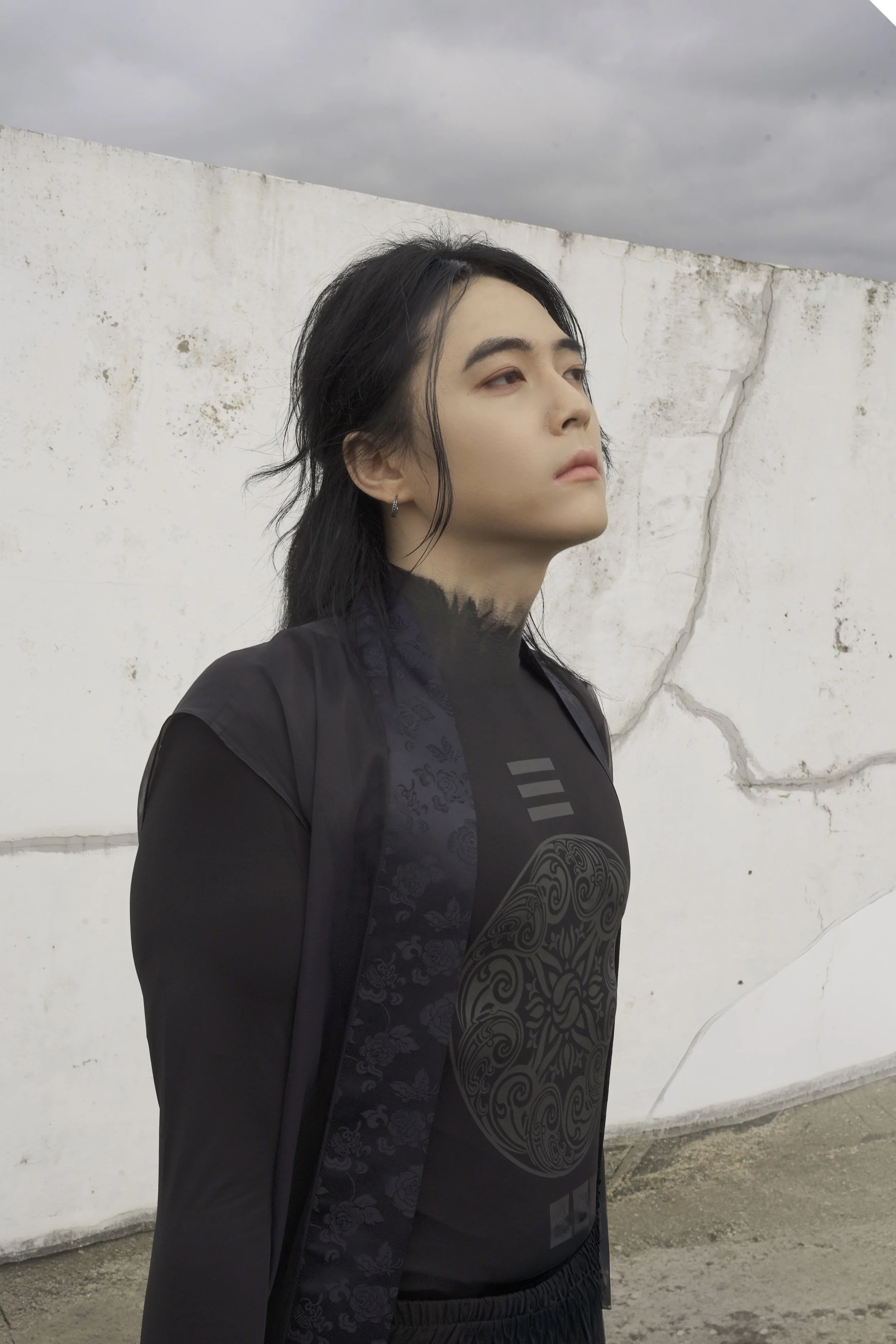 A woman with black hair and earrings standing outdoors against a white wall with visible cracks and a cloudy sky.