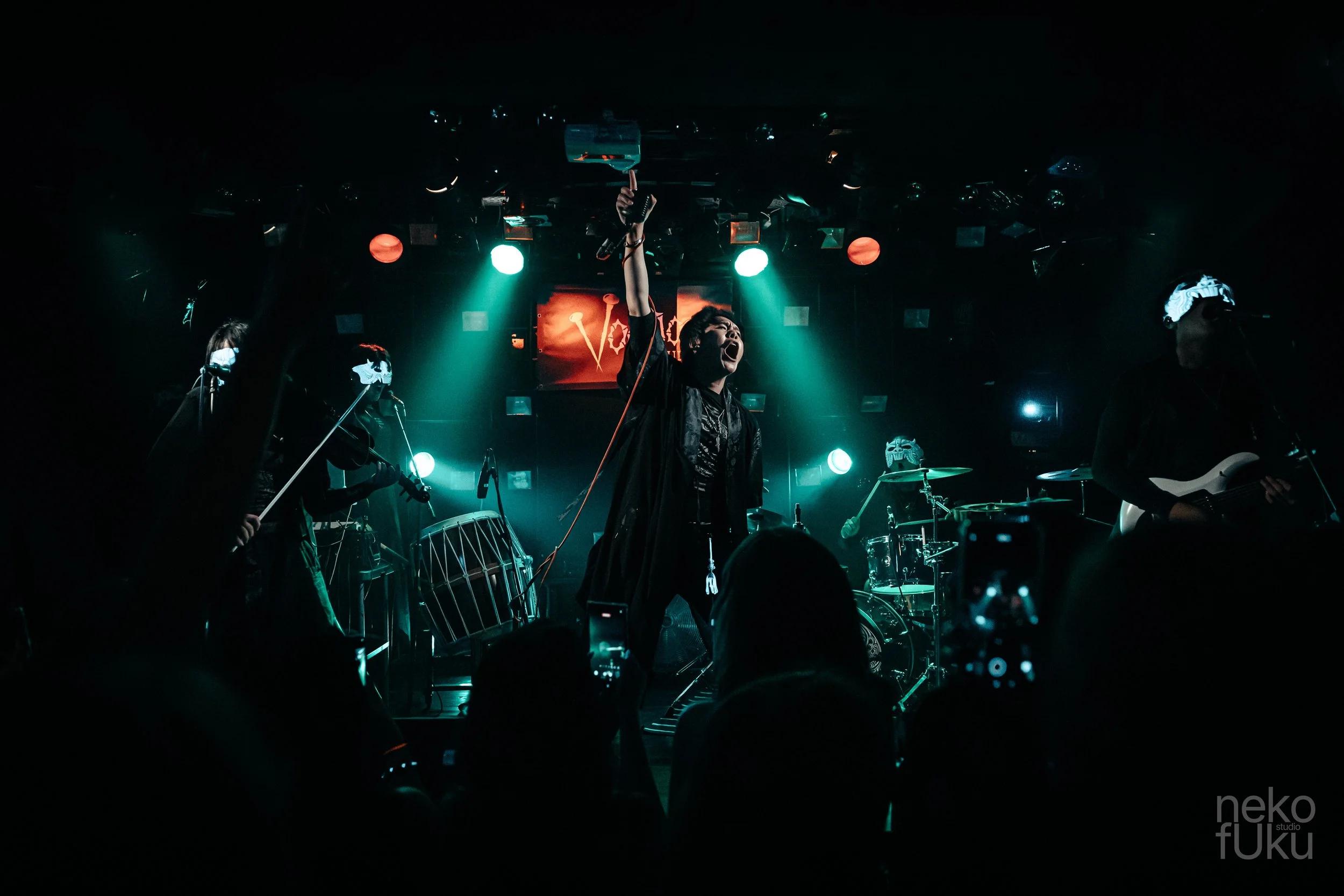 A live music performance on stage with a singer holding a microphone, surrounded by band members playing instruments, under colorful stage lights.
