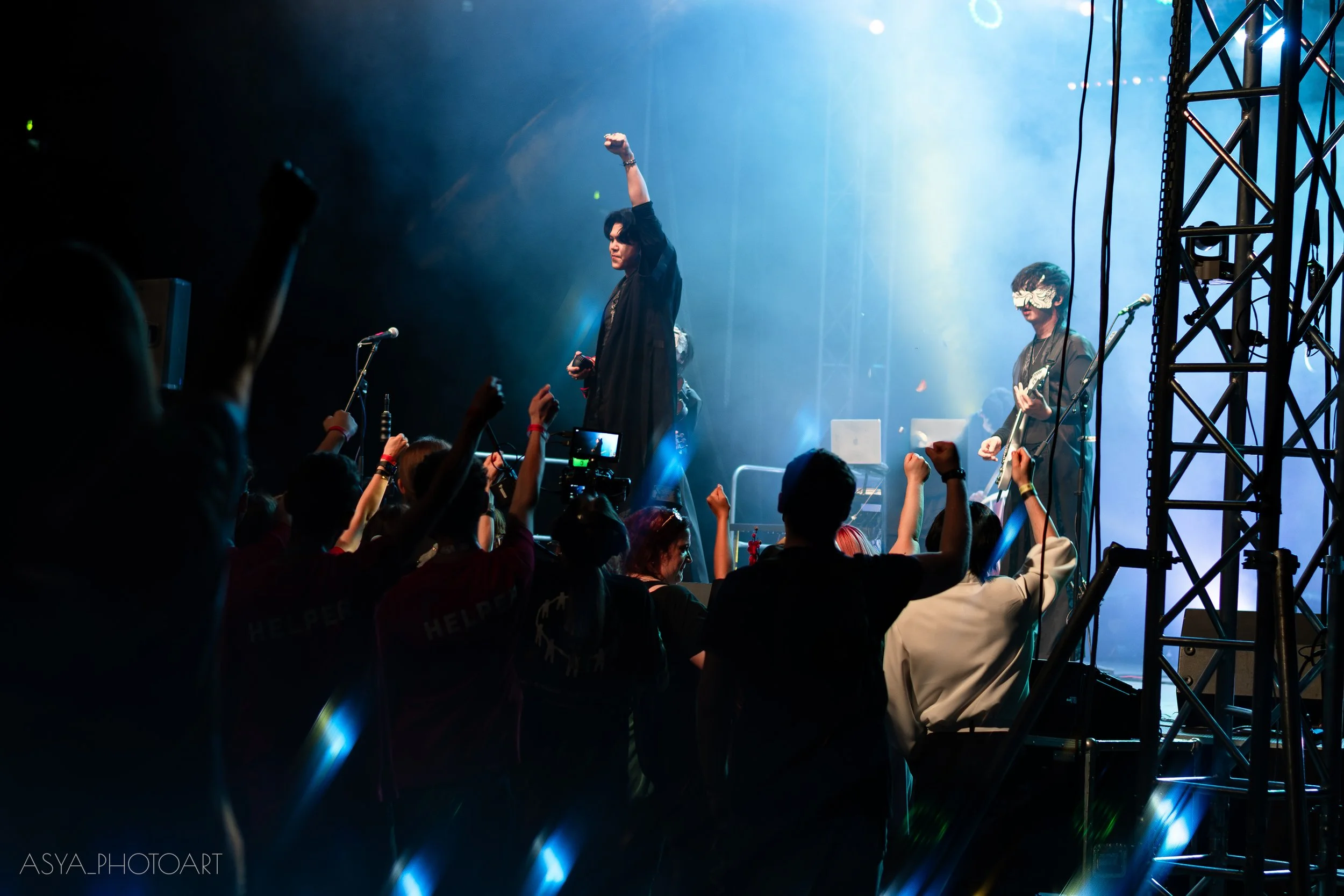 A female singer in black standing on stage with one arm raised, performing at a concert. A guitarist wearing a decorative mask is playing to her right. The audience in front has their hands raised, capturing the moment with phones. Stage lighting cre
