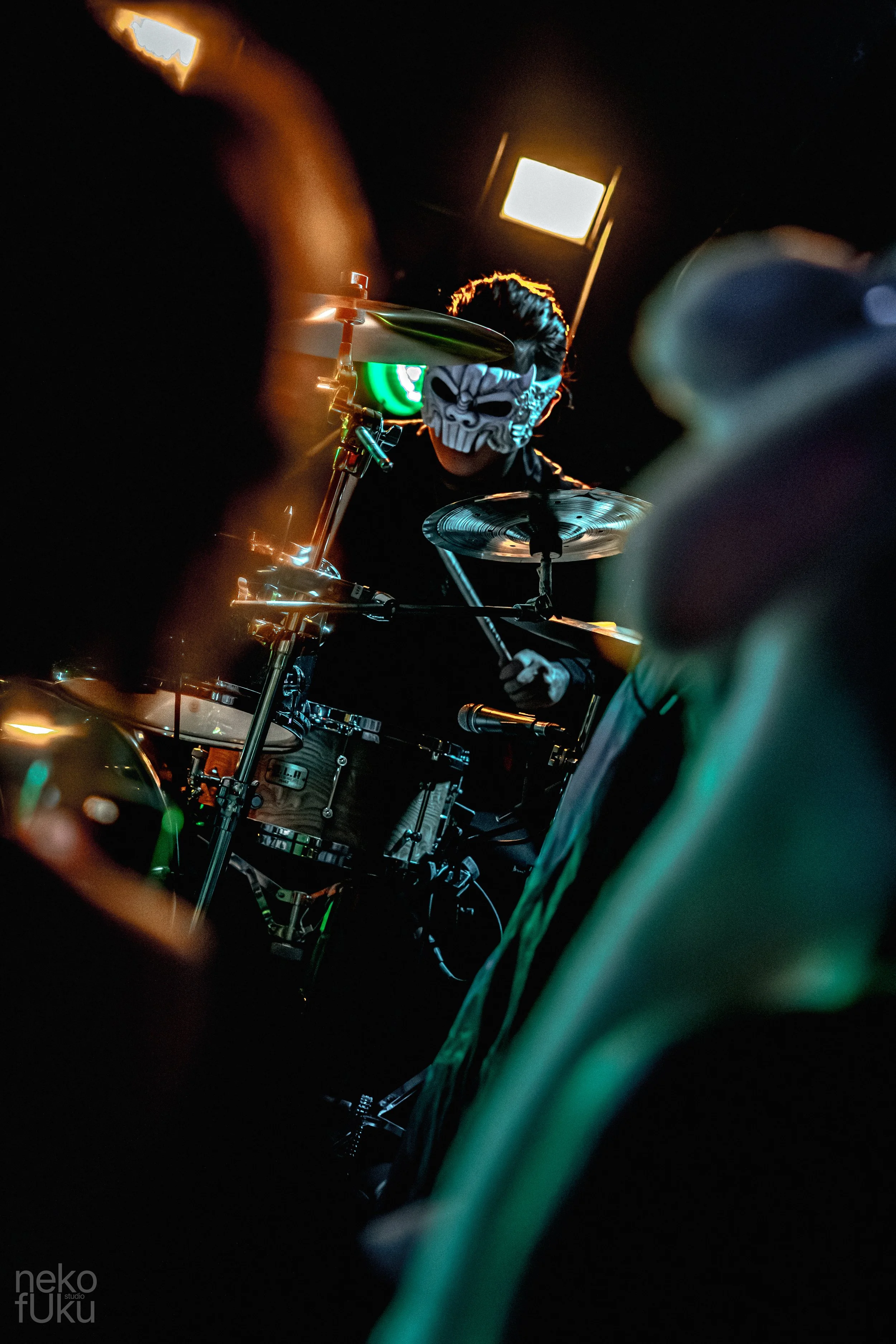 Person wearing a white skull mask playing drums in a dark setting with some bright lights in the background.