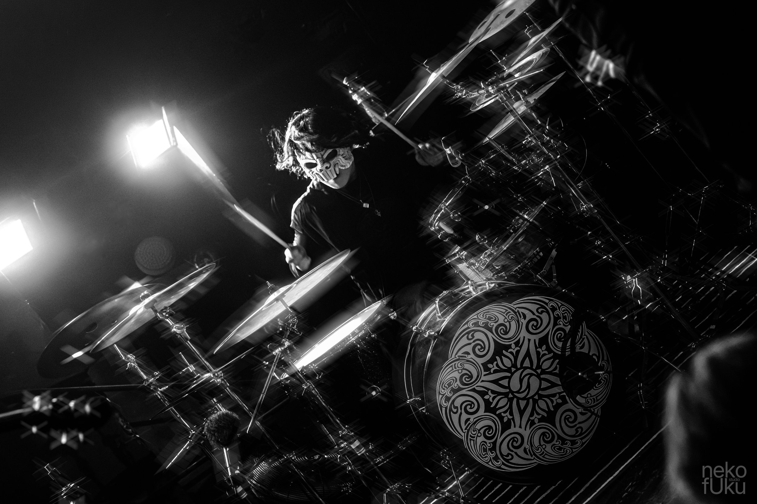 A musician wearing a mask is playing drums on stage, illuminated by stage lights in black and white.