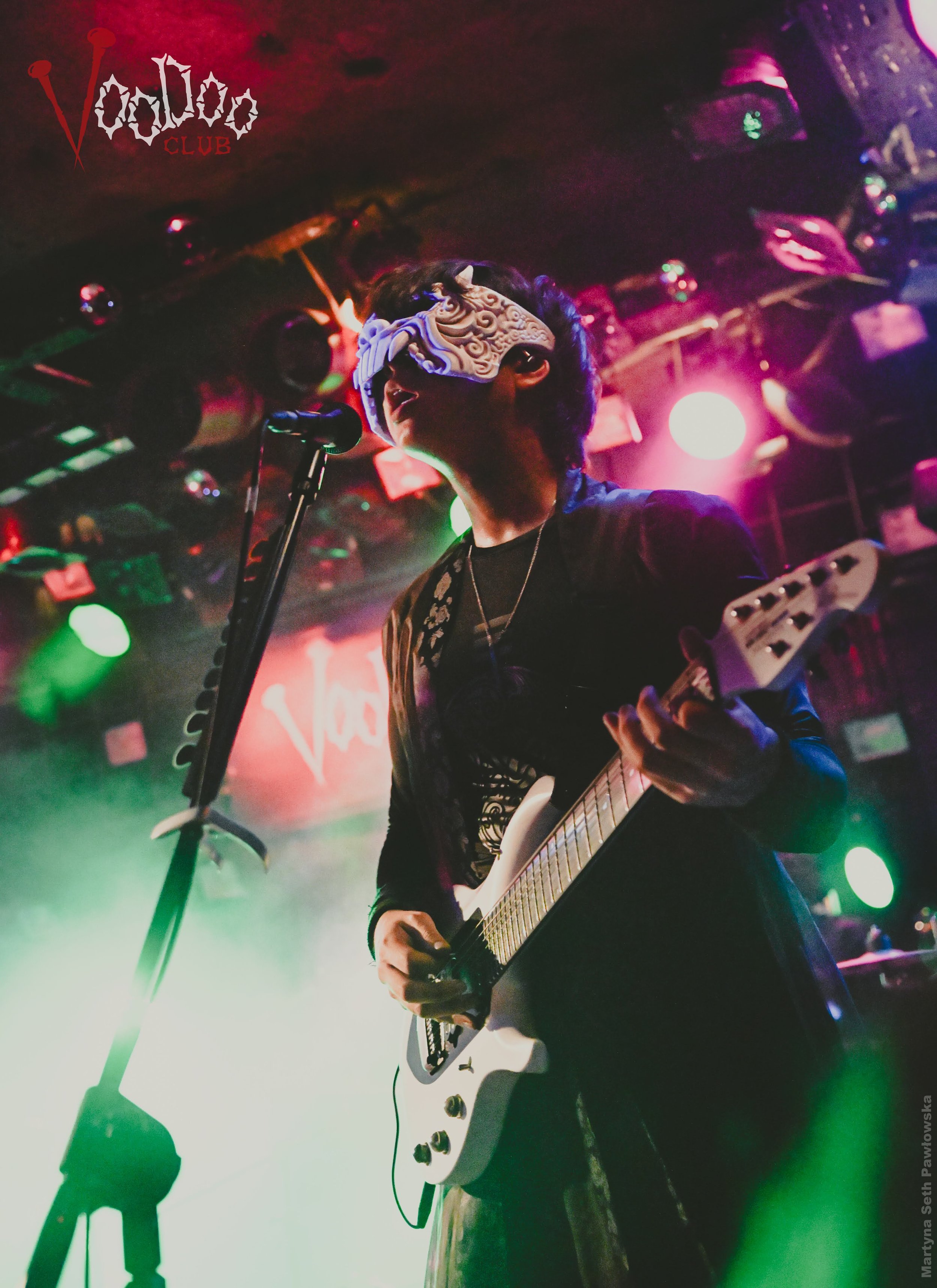 A person wearing a mask and playing an electric guitar on stage with colorful lights. There is a microphone in front of them. The background features a sign that reads 'Volda CLUB' with a stylized logo.