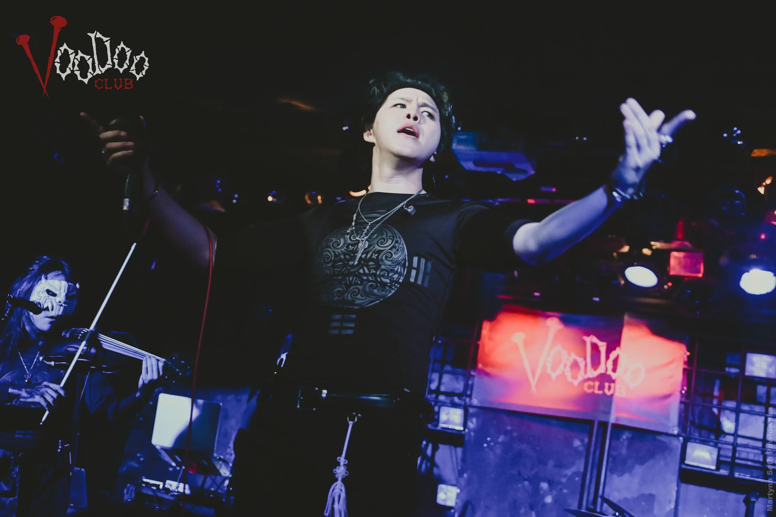 A woman with short black hair singing in a dark nightclub with purple and orange lighting, wearing a black shirt with a largest intricate circular design, multiple necklaces, and has her arms raised. There are other people, including a person with lo