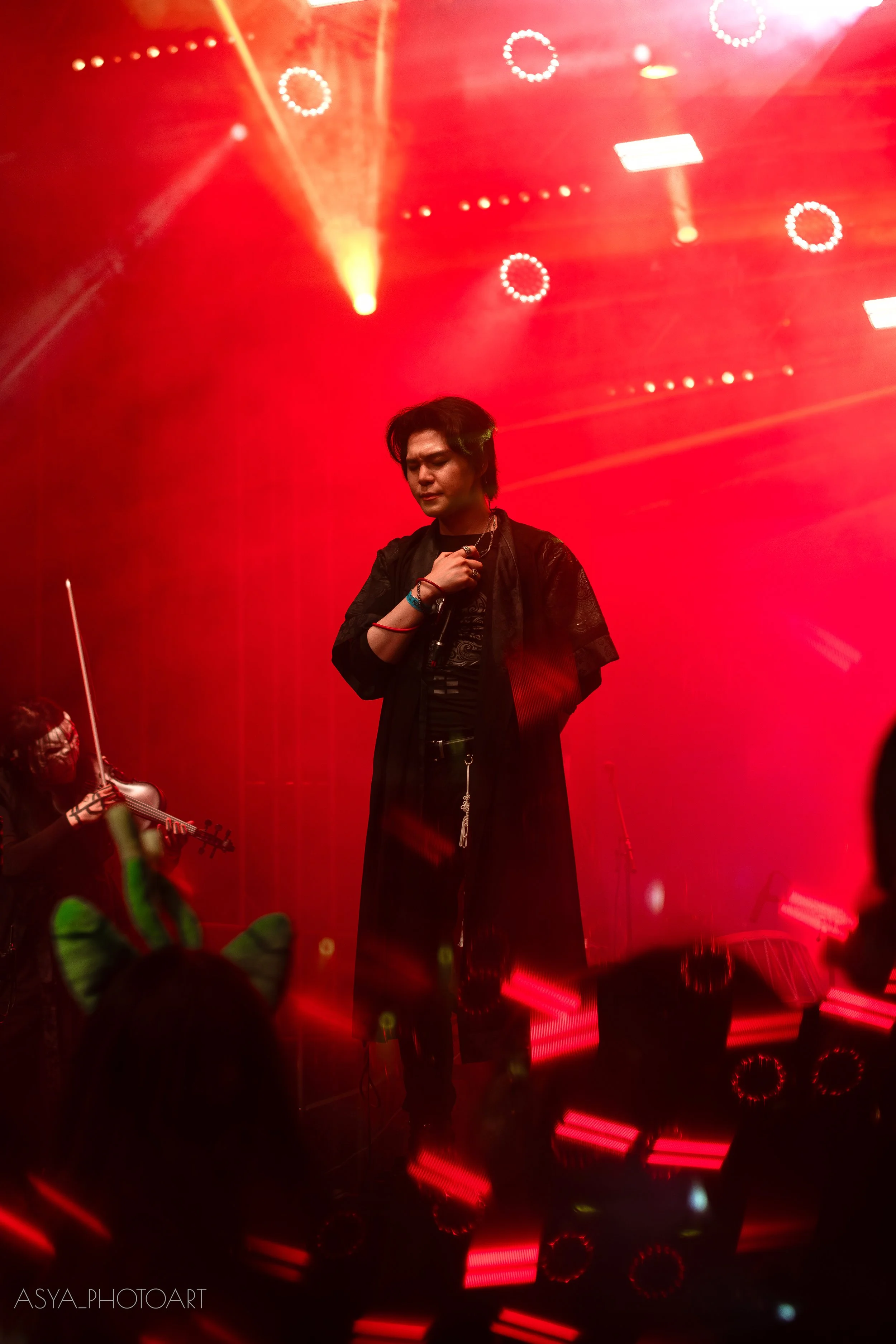 A male singer with black hair performing on a stage with red lighting and circular light fixtures, holding a microphone with his eyes closed.