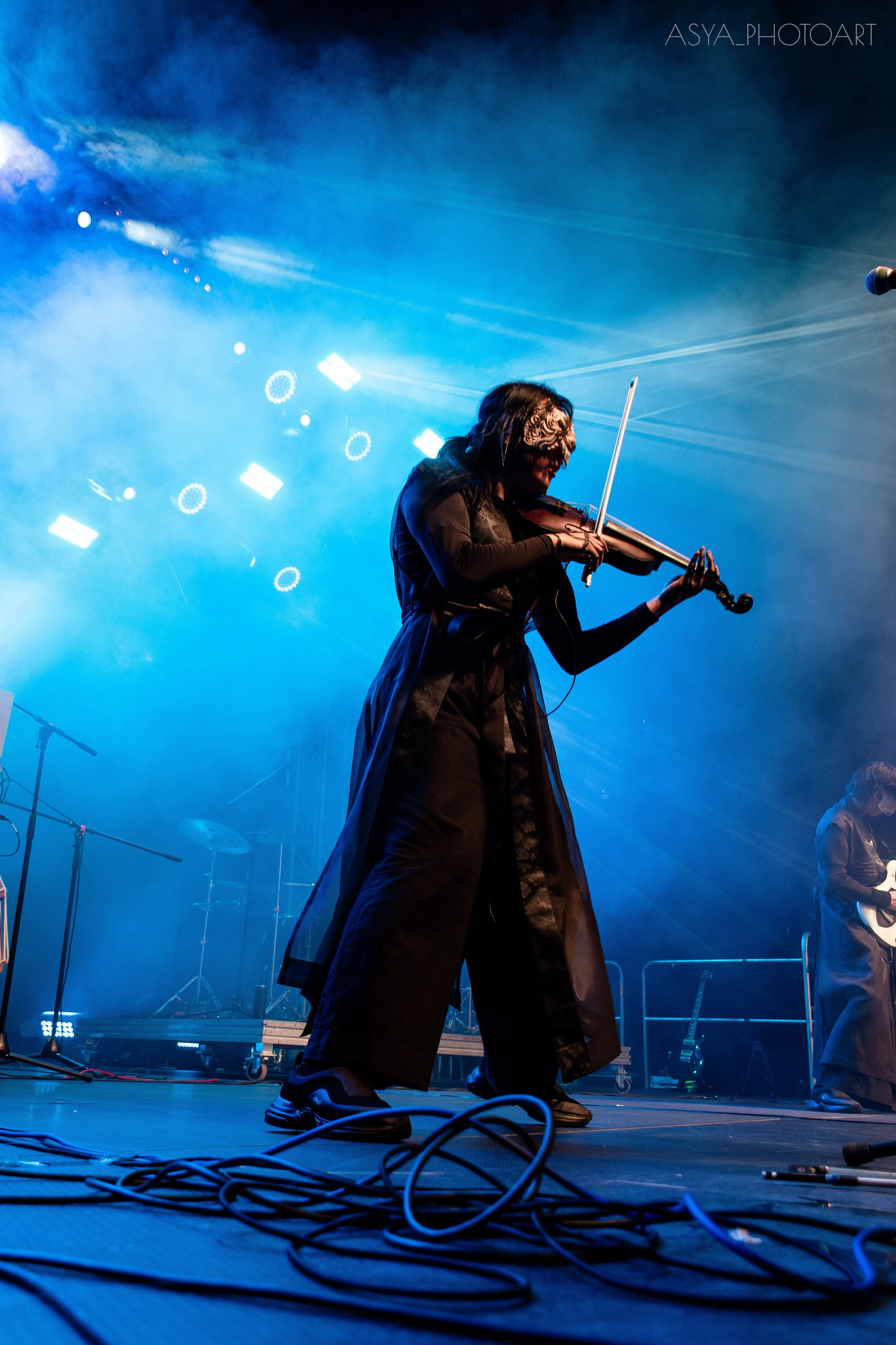 A musician wearing a mask plays a violin on stage illuminated by blue stage lights, with cables on the floor and instruments in the background.