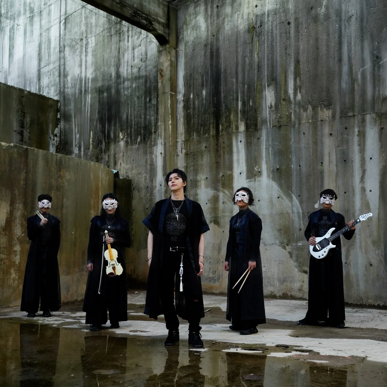 Group of five individuals standing in an abandoned, industrial-looking area with tall concrete walls, wearing black clothing and white mask, some holding musical instruments including a guitar and a violin.