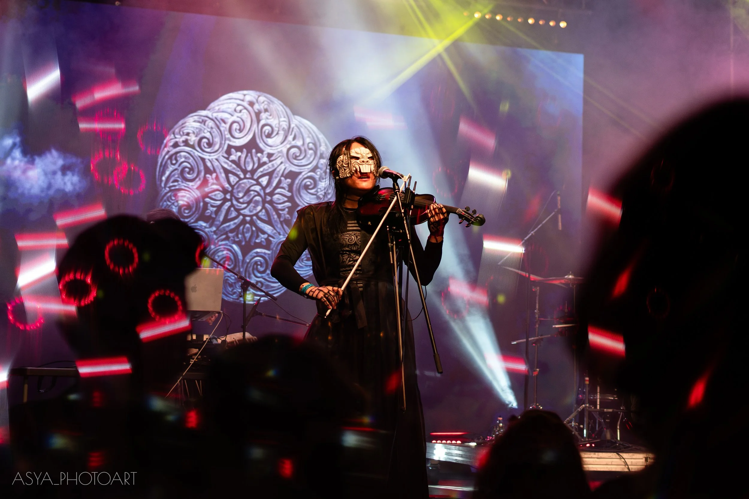 A performer wearing a decorative mask with swirling patterns playing a violin on stage, illuminated by colorful stage lights with a large, ornate background display.