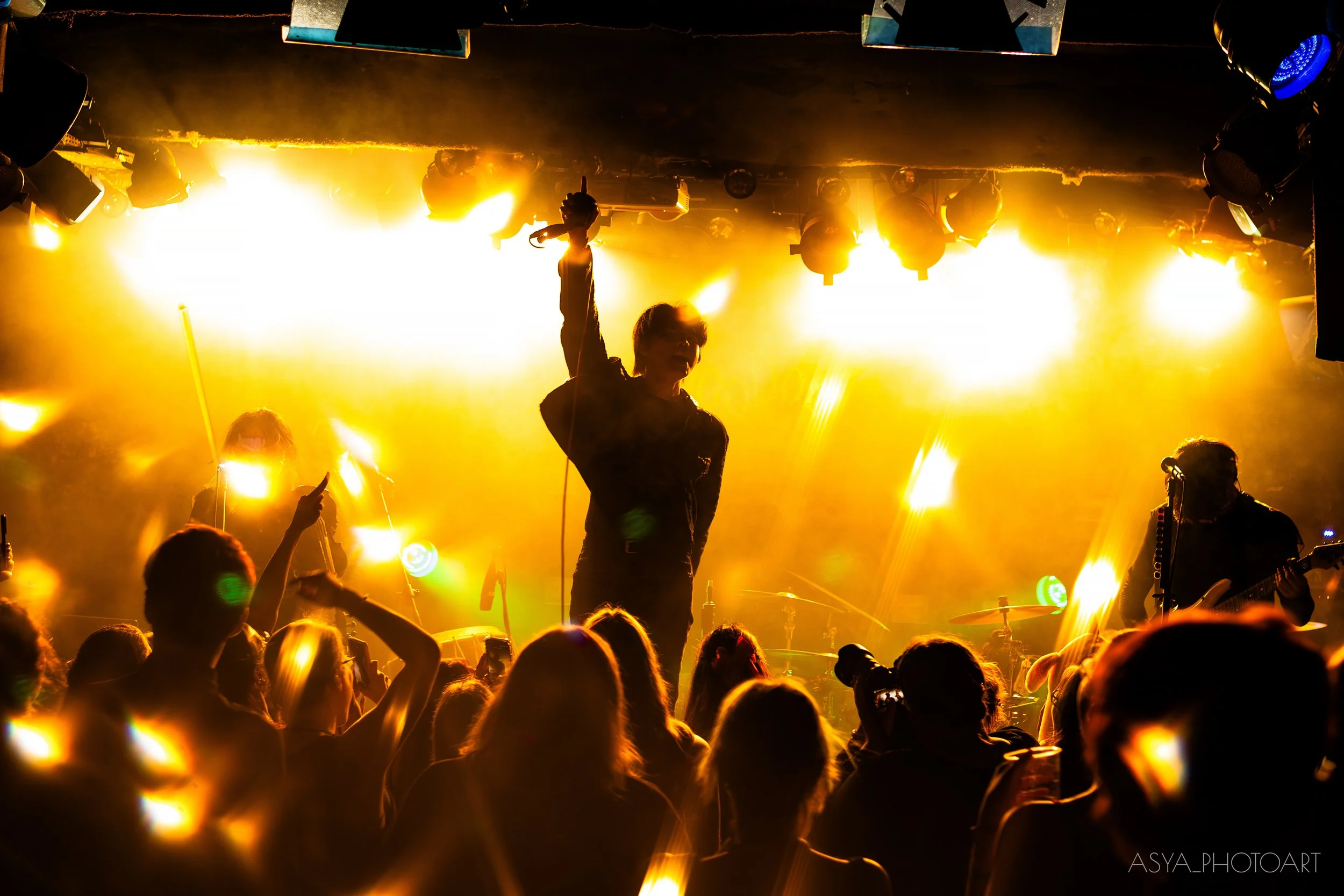 A live concert scene with a singer and band performing on stage, illuminated by bright yellow and orange lights, with an audience in the foreground capturing the performance.