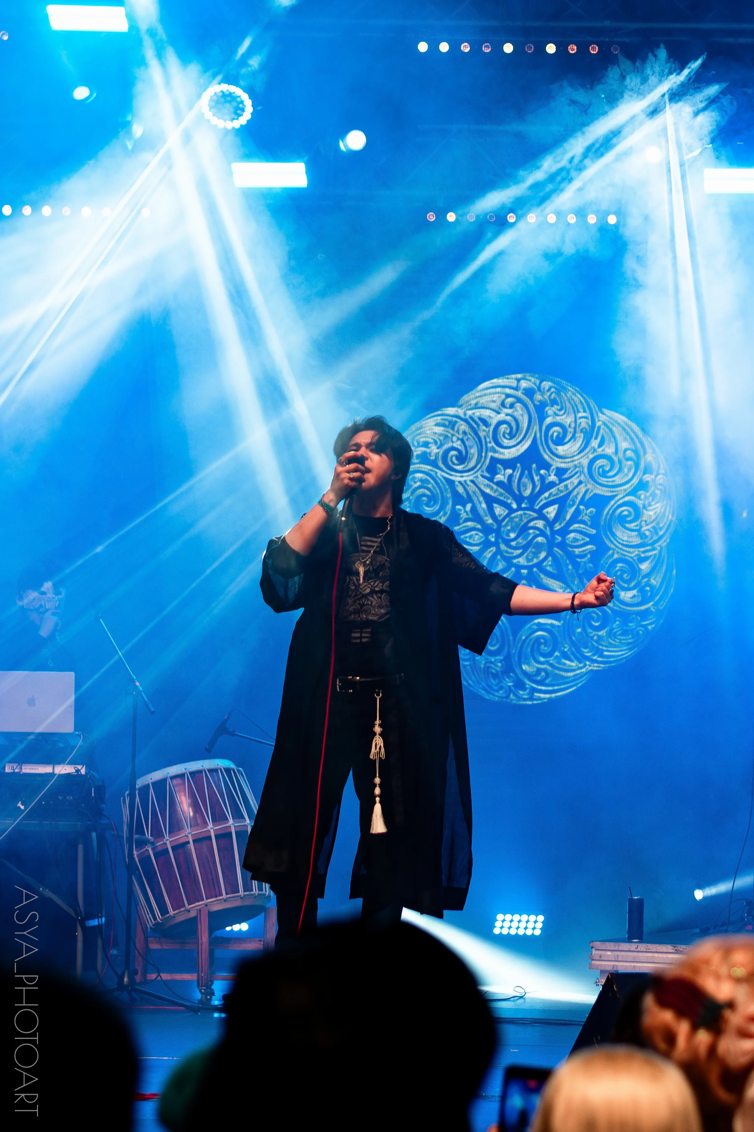 A singer performing on stage with blue lighting and decorative background, surrounded by musical instruments and equipment.