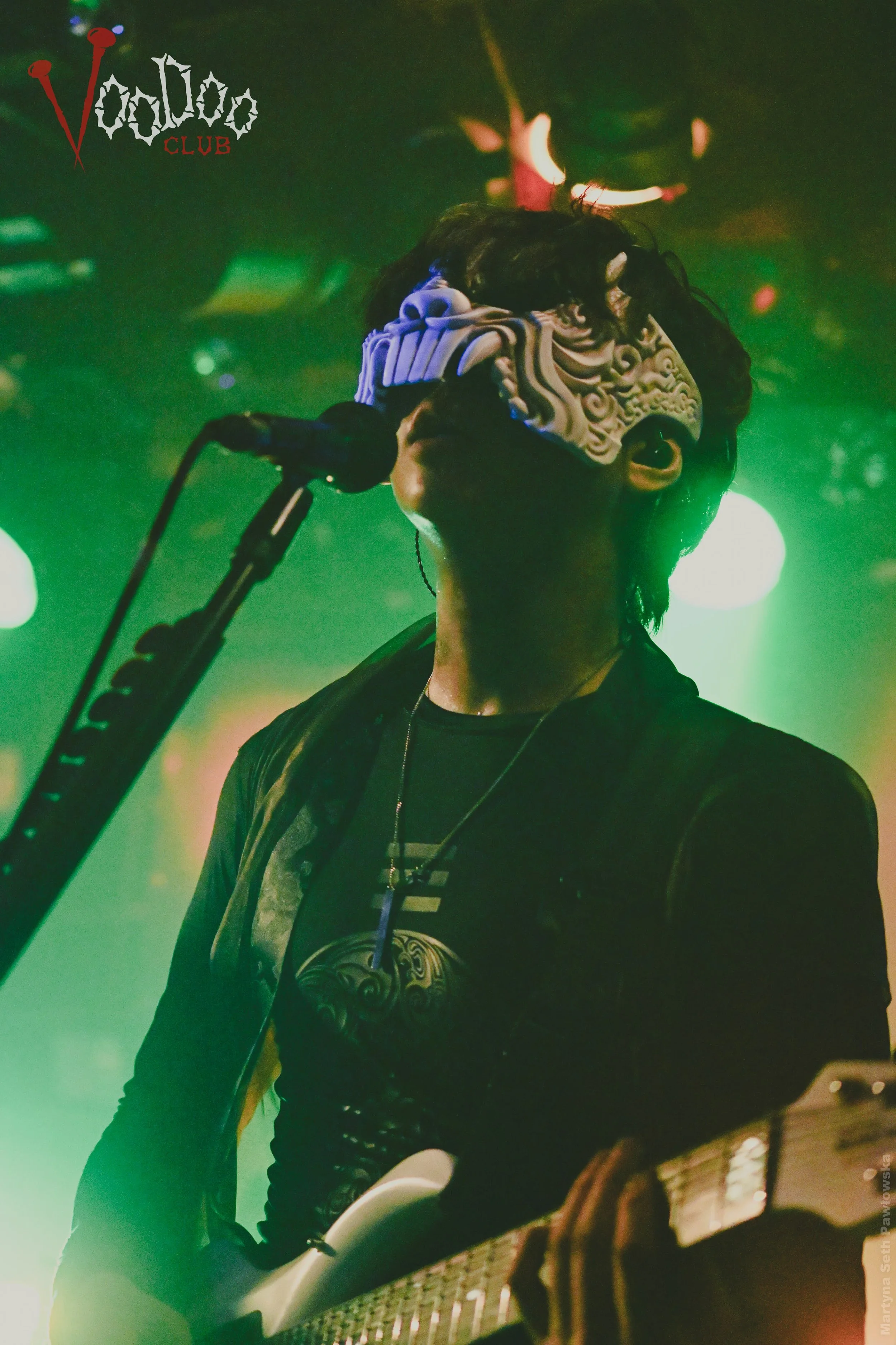A person wearing a mask with intricate patterns and designs playing an electric guitar on stage with colorful lighting and a brick wall in the background, at a club named 'Voodoo Club'.