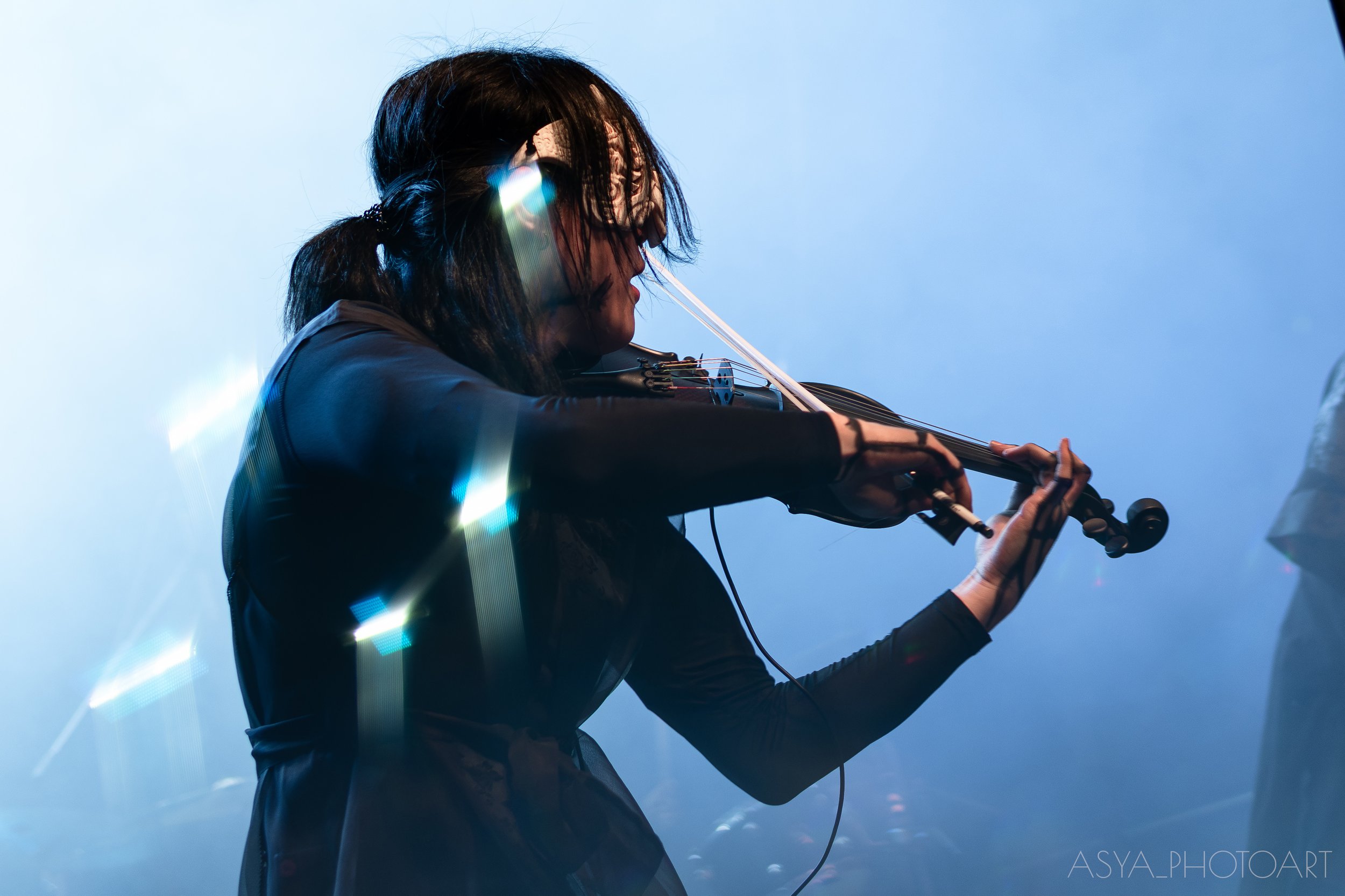 A woman with dark hair playing the violin against a blue sky background, illuminated by light effects.