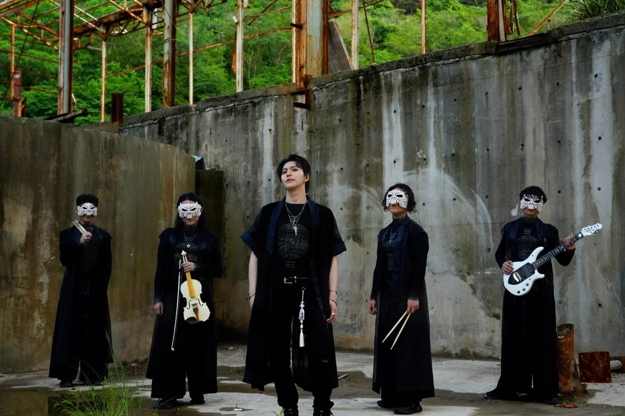 Five individuals dressed in black with white masks, some holding musical instruments like a violin and an electric guitar, standing against a weathered concrete wall in an outdoor setting with green trees in the background.