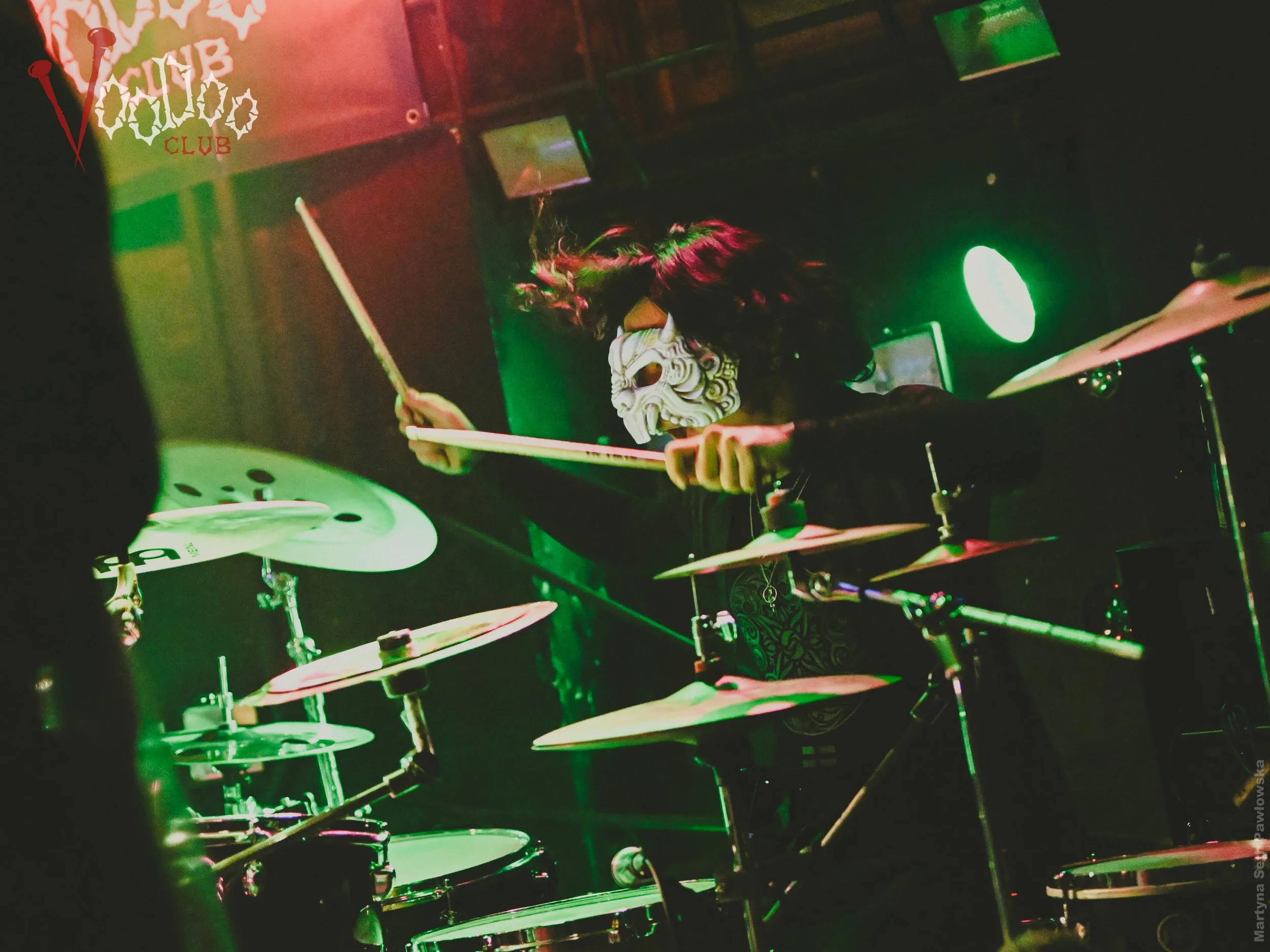 A person wearing a skeleton mask playing drums at a club with neon green and pink lighting.