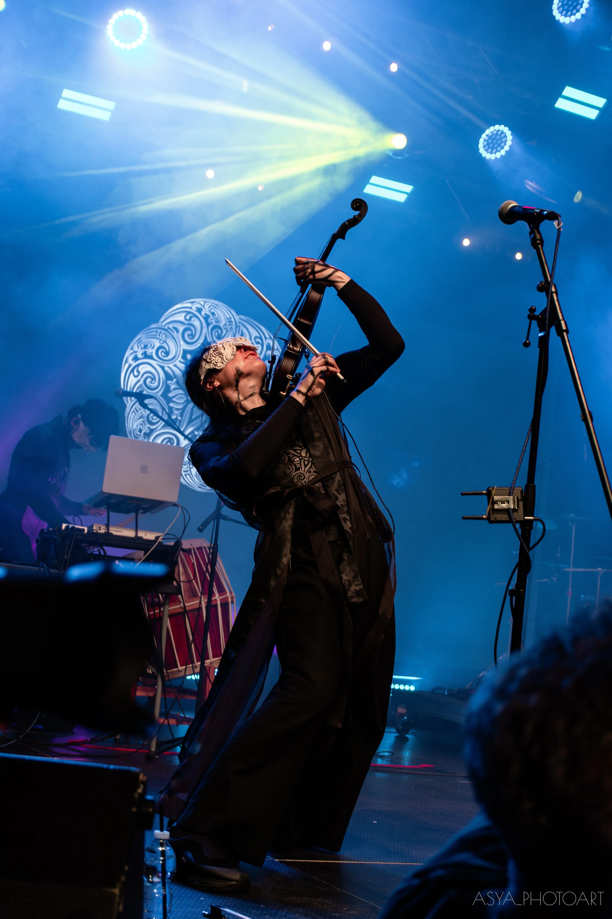 A musician playing a violin on stage with colorful lights and fog effects, with electronic equipment and a person working at a laptop in the background.