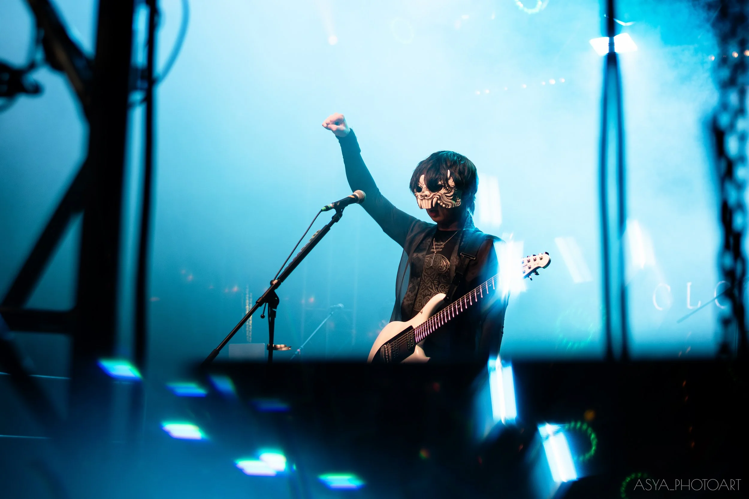 A person performing on stage, wearing a skull mask and playing an electric guitar, with blue stage lighting.