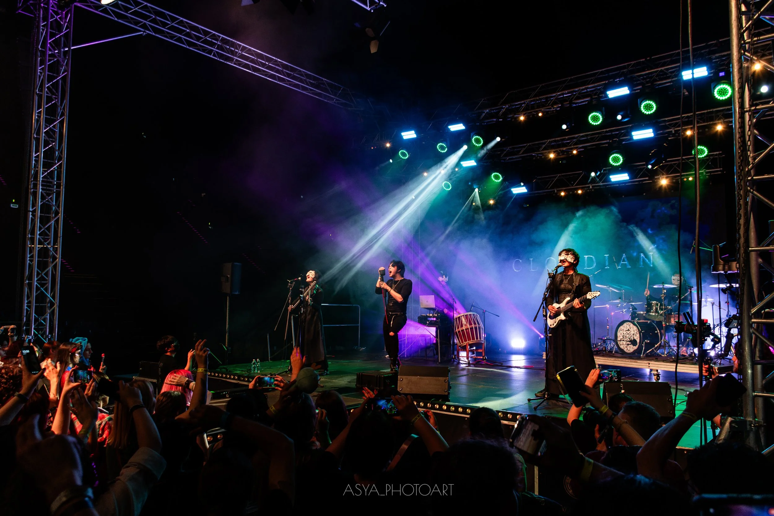 A live band performing on stage with colorful stage lights and smoke effects, audience members taking photos.
