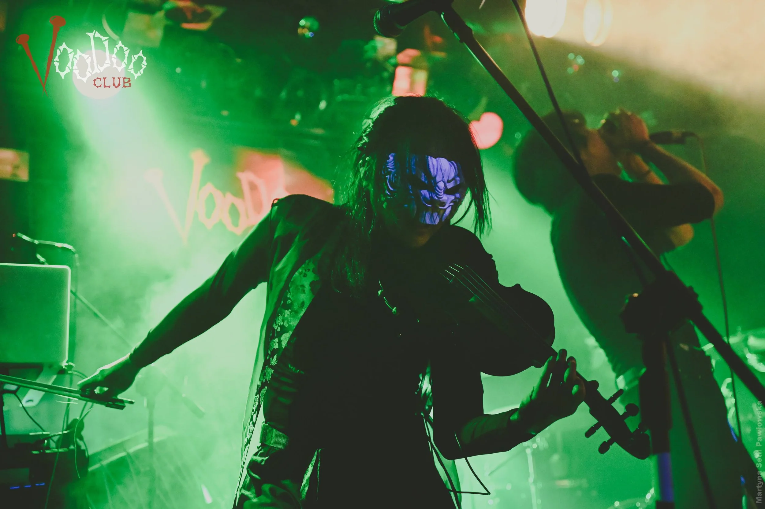 A live music performance at a nightclub featuring a musician with long black hair wearing a skull mask, playing an electric guitar under green stage lighting, with a microphone and electronic equipment visible in the background.