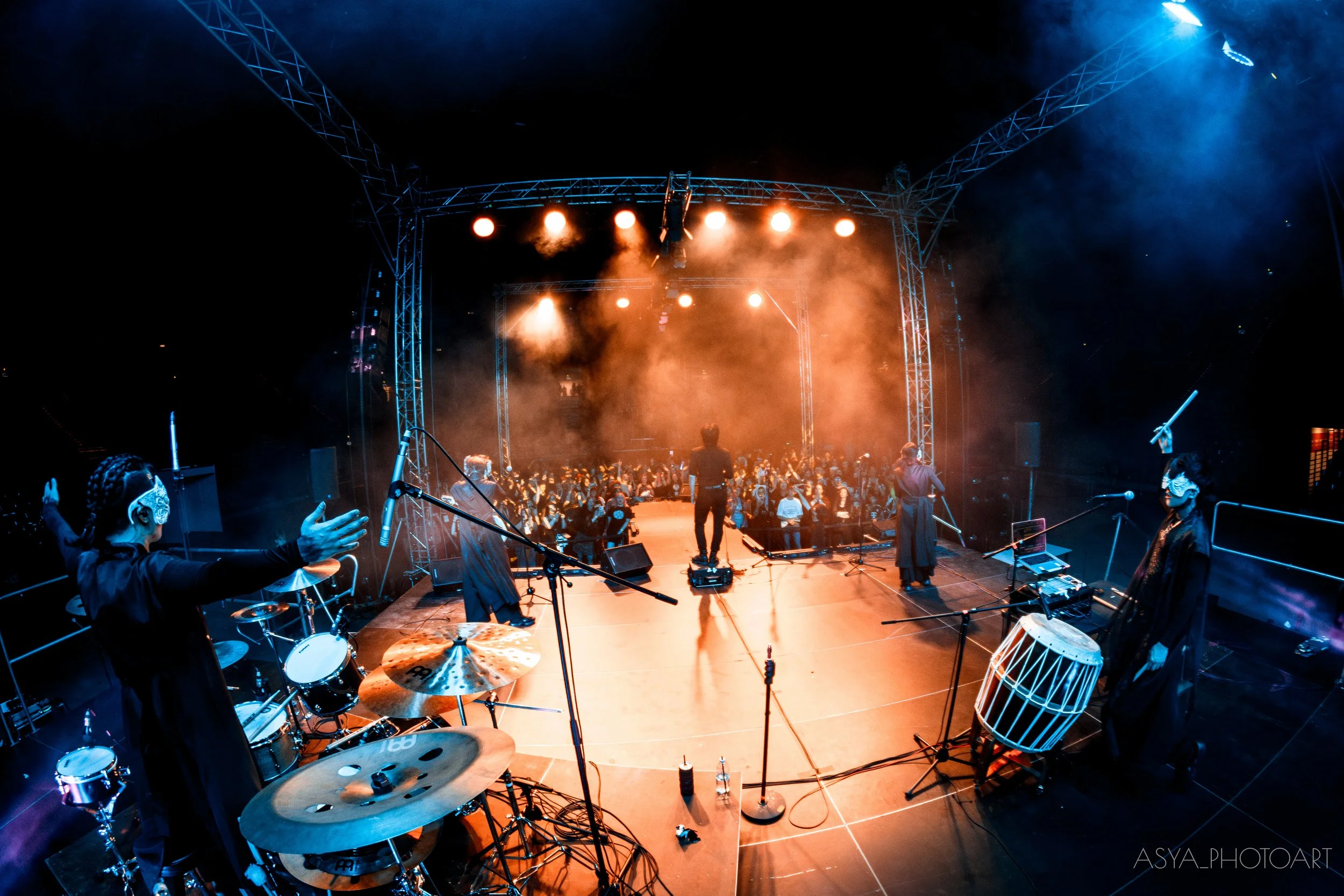 A concert stage with musicians playing, colorful stage lighting, and a large audience in the dark. Musicians include a drummer, percussionist, and others, with equipment and instruments visible.