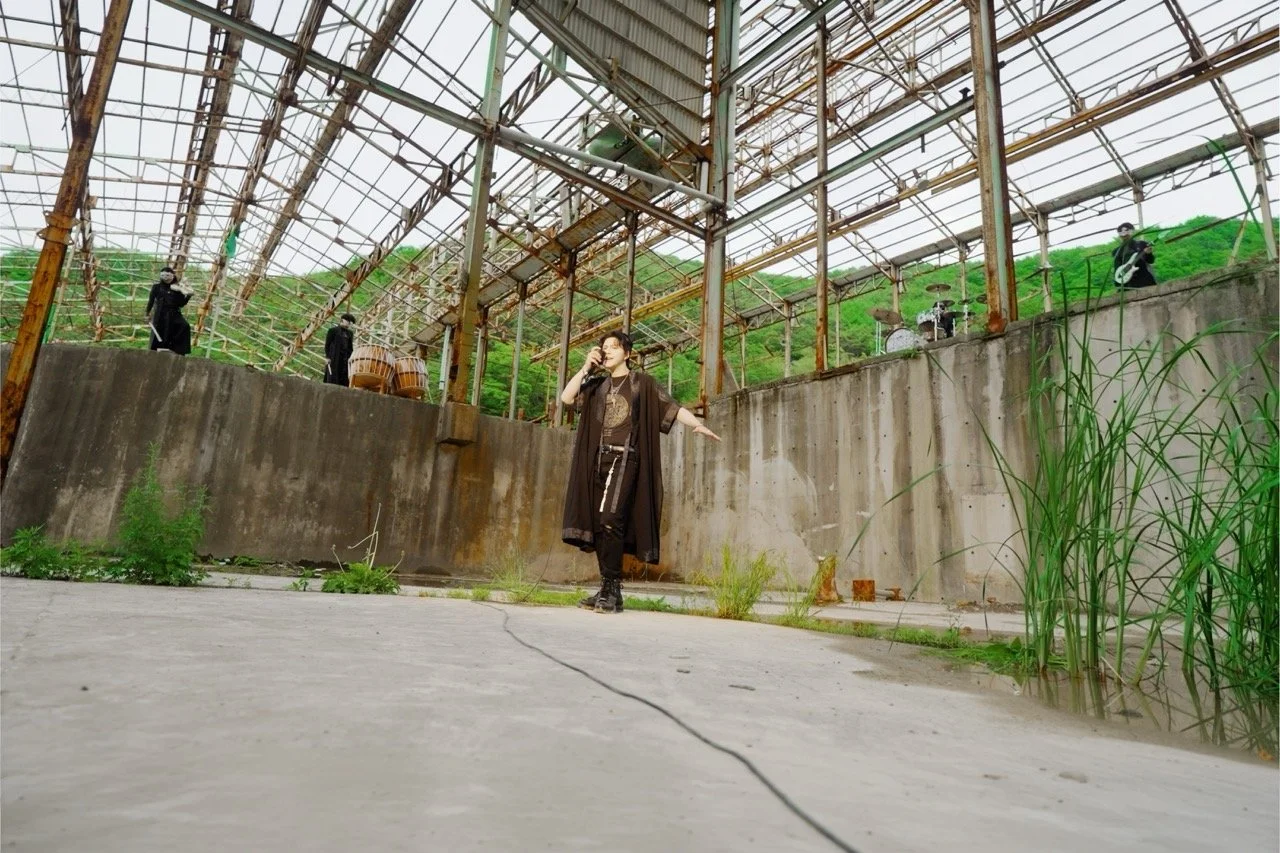 A woman singing into a microphone in an abandoned industrial building with musicians playing instruments on the platform behind her.