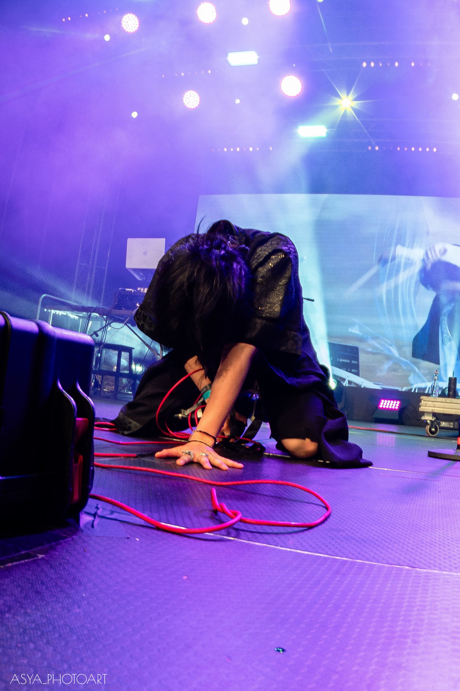 A person is kneeling on stage, bent forward with their head down, surrounded by stage equipment, with colorful bright stage lights overhead, and a large screen or backdrop behind them.