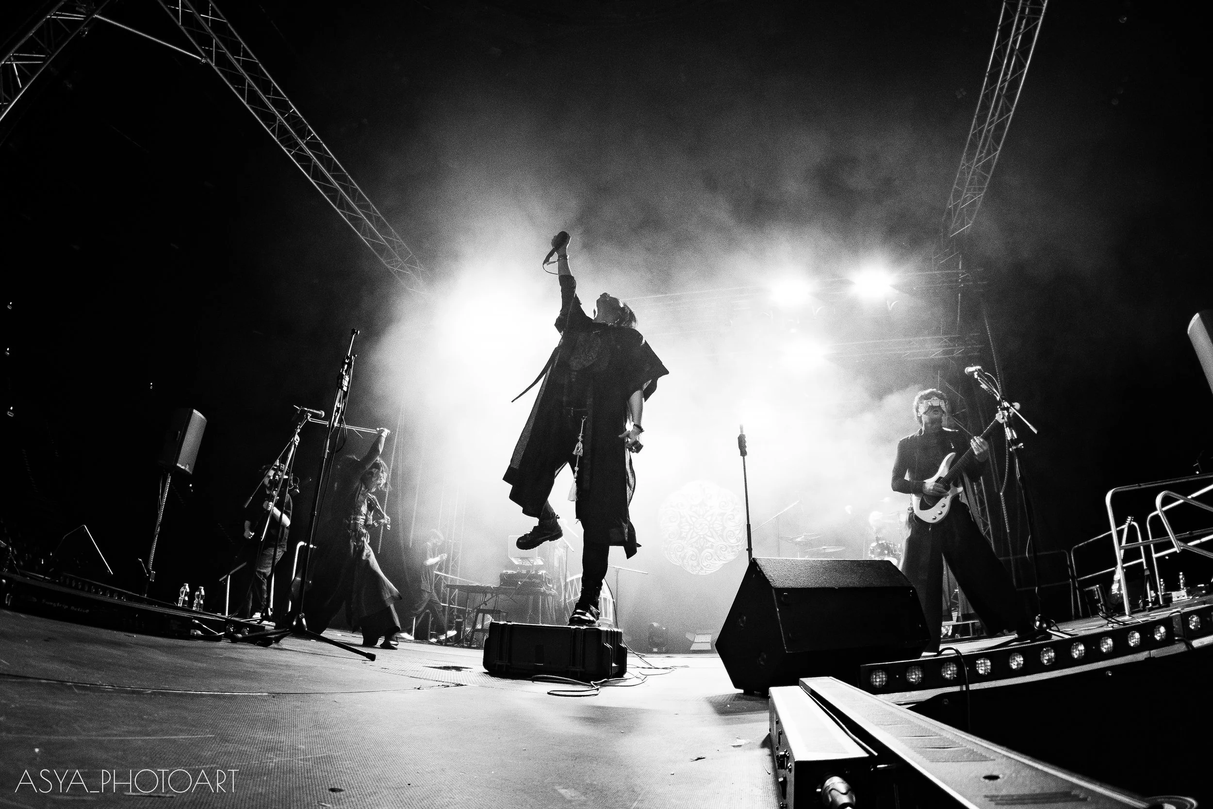 Black and white photo of a person jumping on stage during a live music performance, with band members playing instruments in the background, stage lighting creating a dramatic effect.