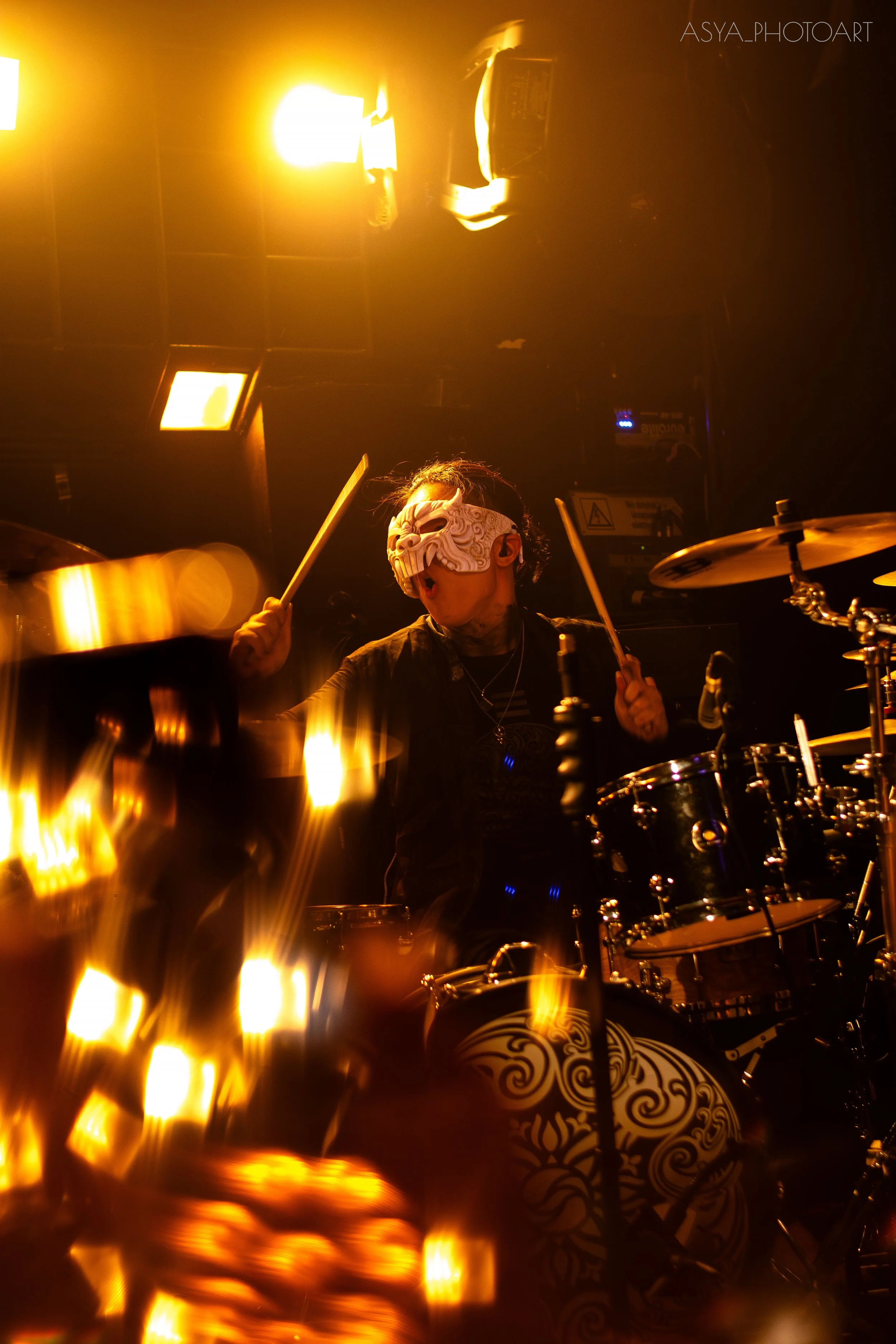 A person playing drums on stage, wearing a white ornate mask and dark clothing, illuminated by warm yellow lighting.