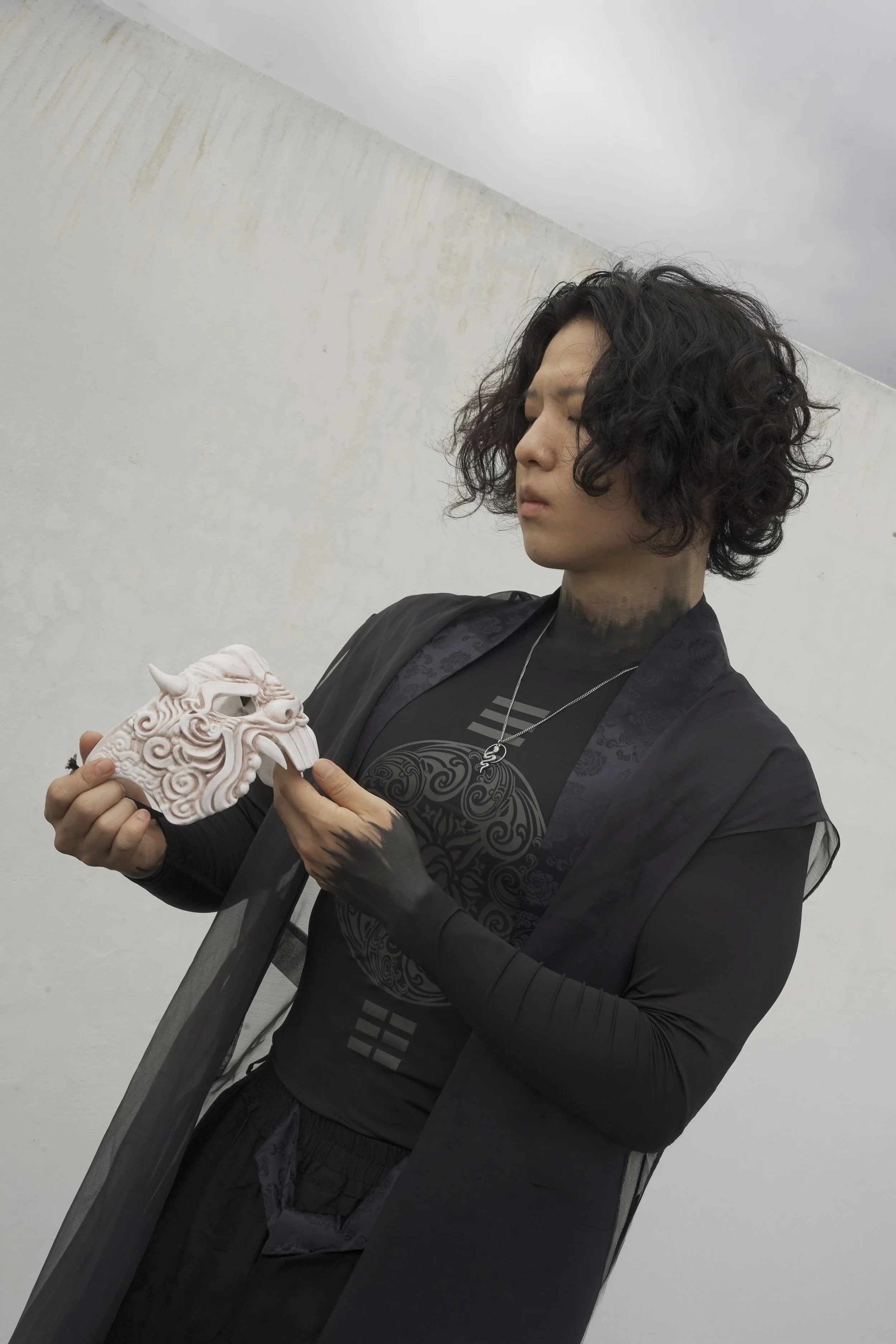 A person with curly black hair and tattoos on their hands holding a detailed dragon-shaped mask or sculpture while standing against a plain white wall.